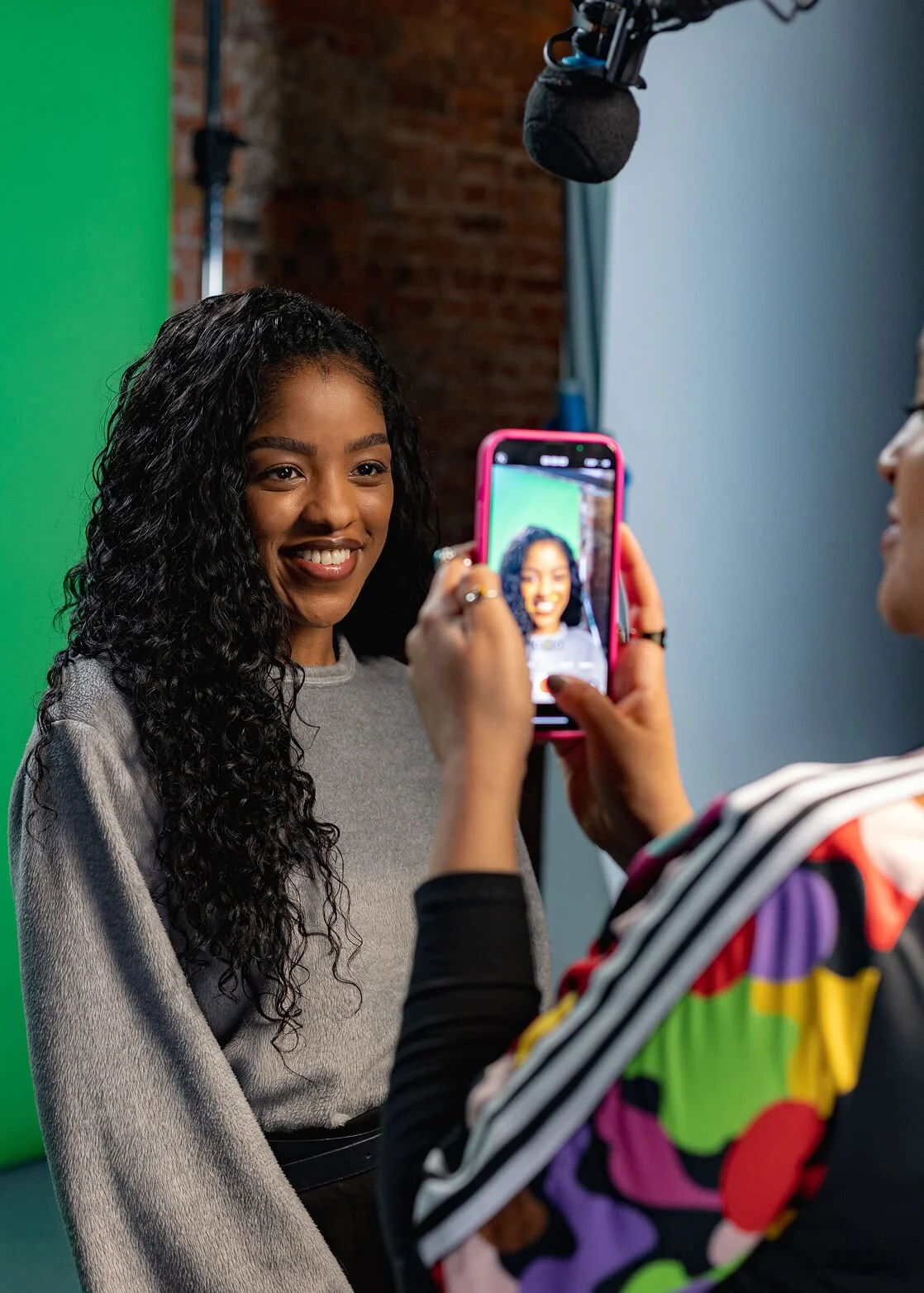 A woman with long curly black hair smiling as she is being photographed with a smartphone by another person.