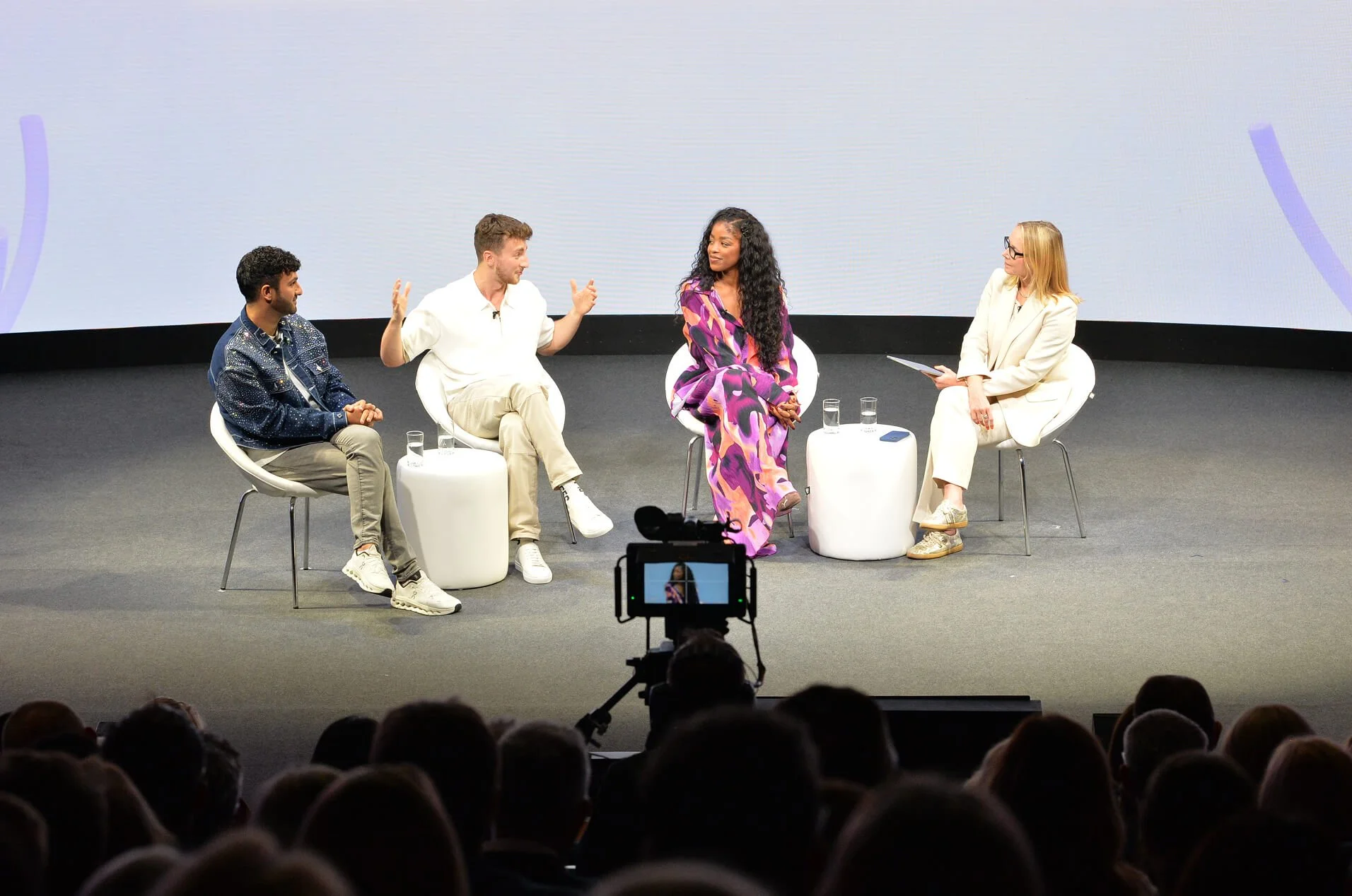 Four people seated on stage, engaged in conversation, with an audience in front of them and a camera recording the event.