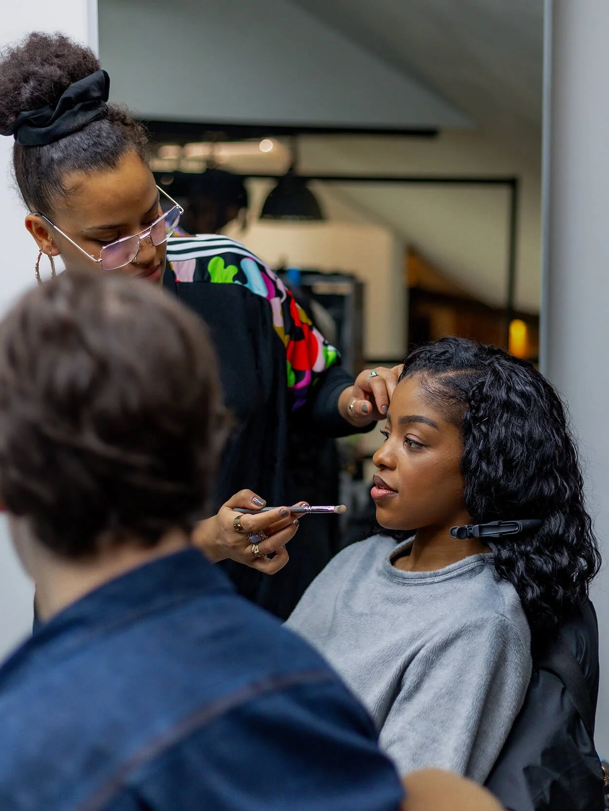 Makeup artist applying makeup to a woman seated in a salon chair while a person with short hair looks on.