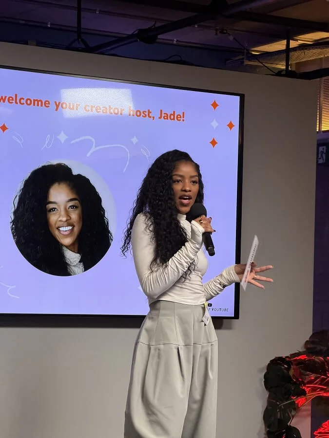 A young woman with long curly hair, wearing a beige top and light-colored pants, is speaking into a microphone in front of a large screen that displays a welcome message for her as the creator host, Jade. The screen shows her photo and colorful decorations.
