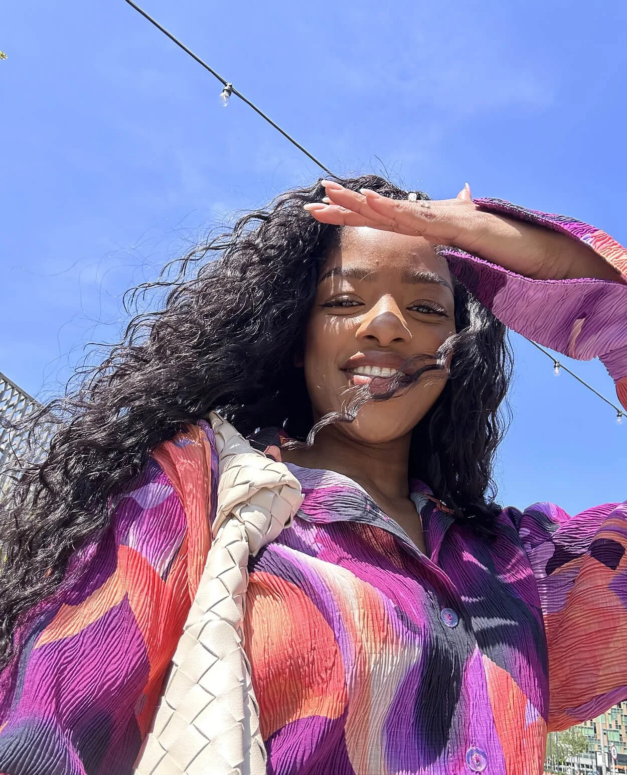 A woman with curly dark hair smiling and shielding her eyes from the sun, wearing a colorful purple, pink, and orange patterned top, carrying a white braided bag over her shoulder, with a bright blue sky and string lights in the background.