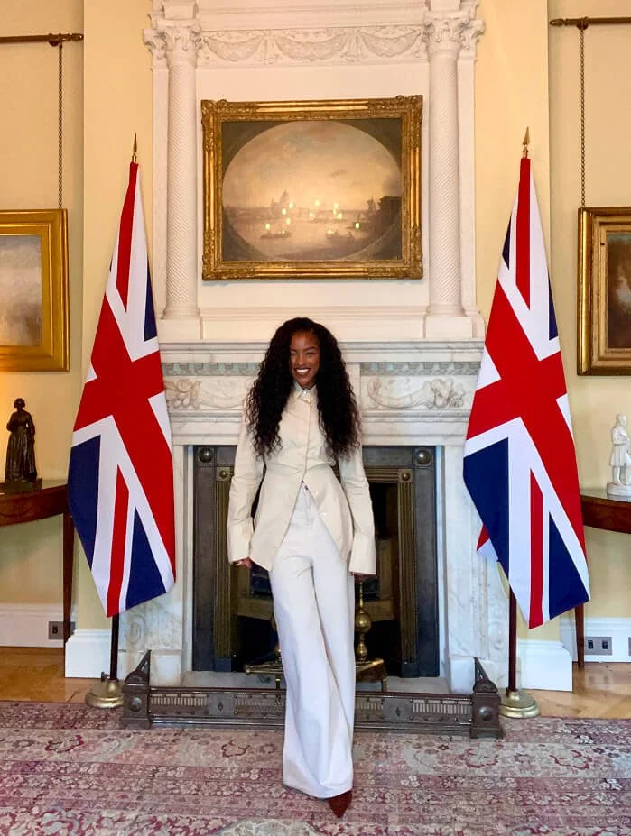 A woman with long curly hair, dressed in a white suit, standing in front of a fireplace with two British flags on each side, inside a room with classic decor.