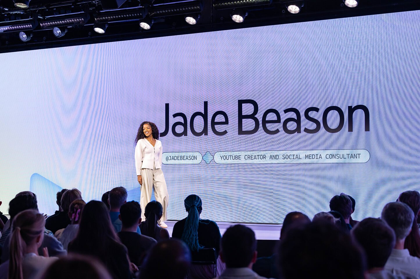 Jade Beason standing on stage during a presentation in front of a large screen displaying her name and social media handle. She is wearing a white outfit and smiling at the audience, which is visible in the foreground.
