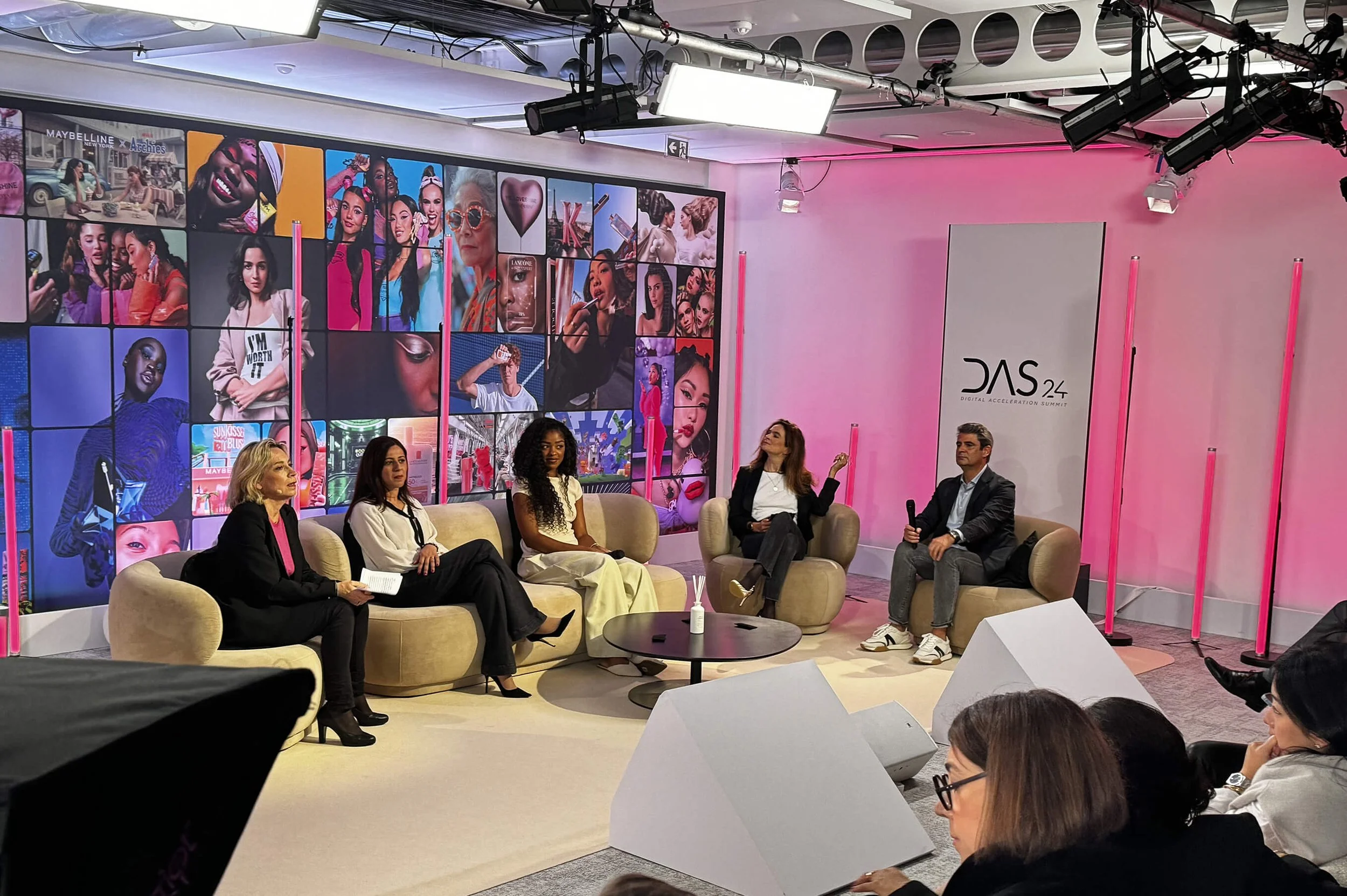 A panel of five women and one man seated on beige couches and chairs, participating in a discussion at the DAS24 Digital Acceleration Summit. The venue features a large wall display with colorful images of fashion, beauty, and pop culture, and has professional lighting and sound equipment.