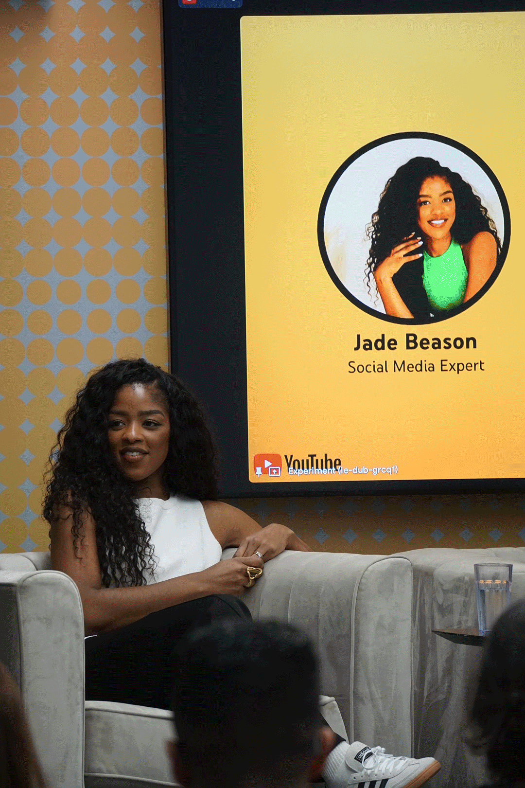 Jade Beason, a woman with curly hair, sitting on a beige couch during a presentation or event, with a large screen behind her displaying her name, photo, and title as a social media expert.