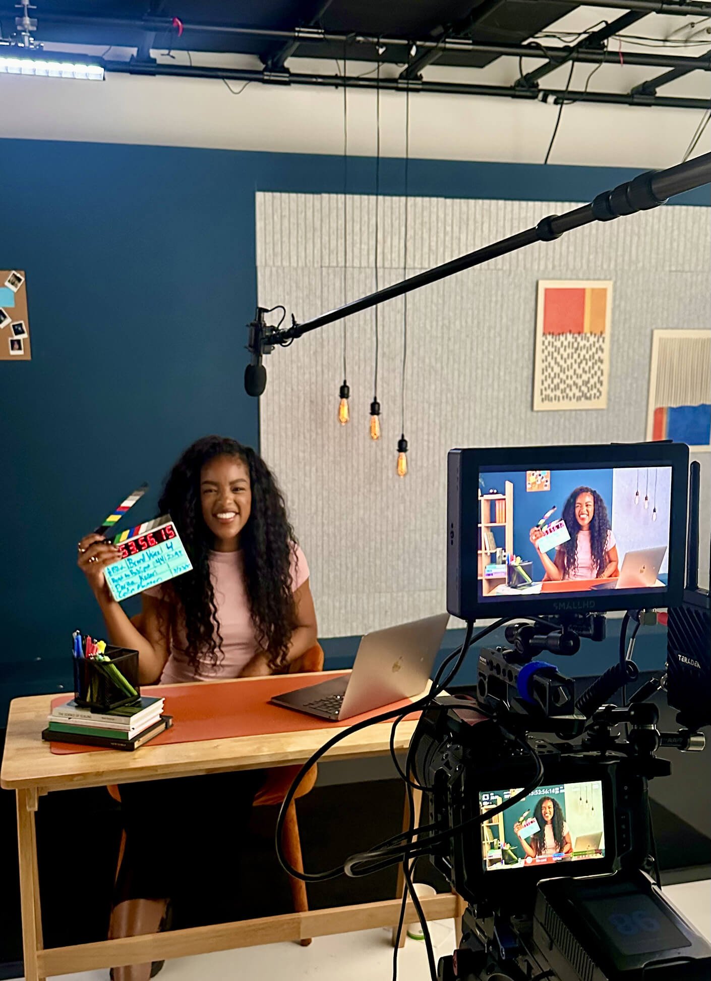 A woman sitting at a desk holding a clapperboard, smiling at the camera. A laptop and a stack of books are on the desk. A camera viewfinder shows her image. The background includes a blue wall with artwork and hanging light bulbs.