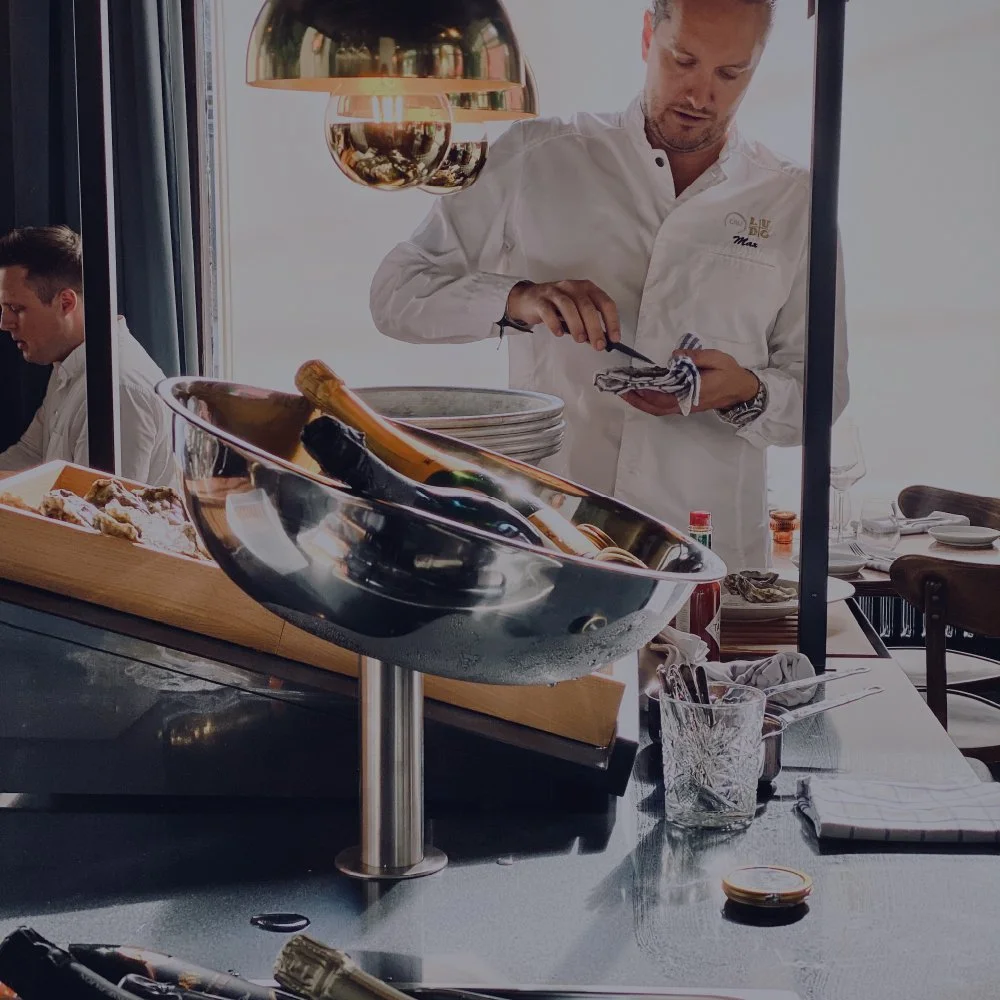 A chef in a white uniform preparing food in a restaurant kitchen, with wine bottles and glasses on the counter and a person in the background.