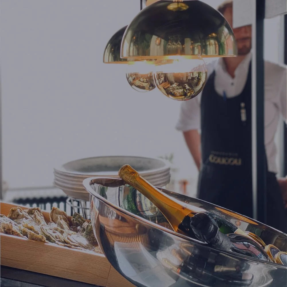 A refrigerator with metallic earrings hanging from the top, a bottle of champagne in a silver ice bucket, and some oysters on a tray in a restaurant setting. A person in a white shirt and black apron is in the background.