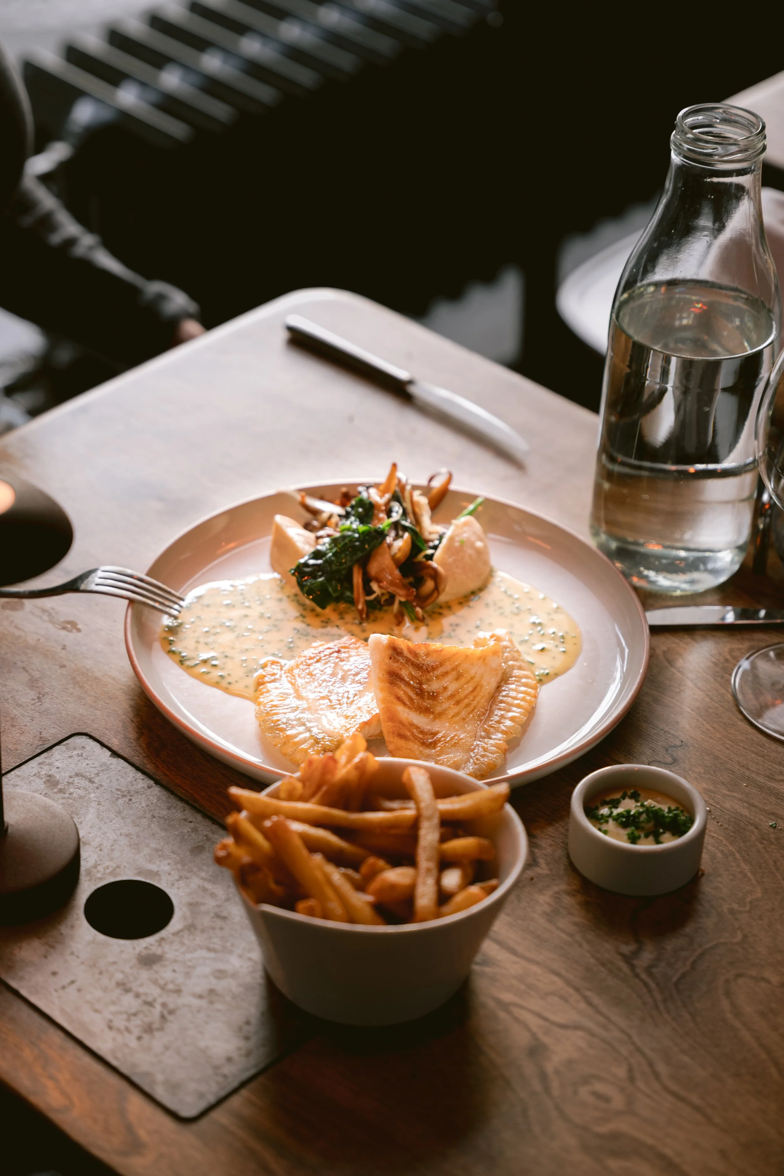 A meal on a wooden table featuring a plate with fish, sauce, and vegetables, a bowl of French fries, a small dish of dipping sauce, a clear glass bottle of water, and a small jar of herbs or seasoning.