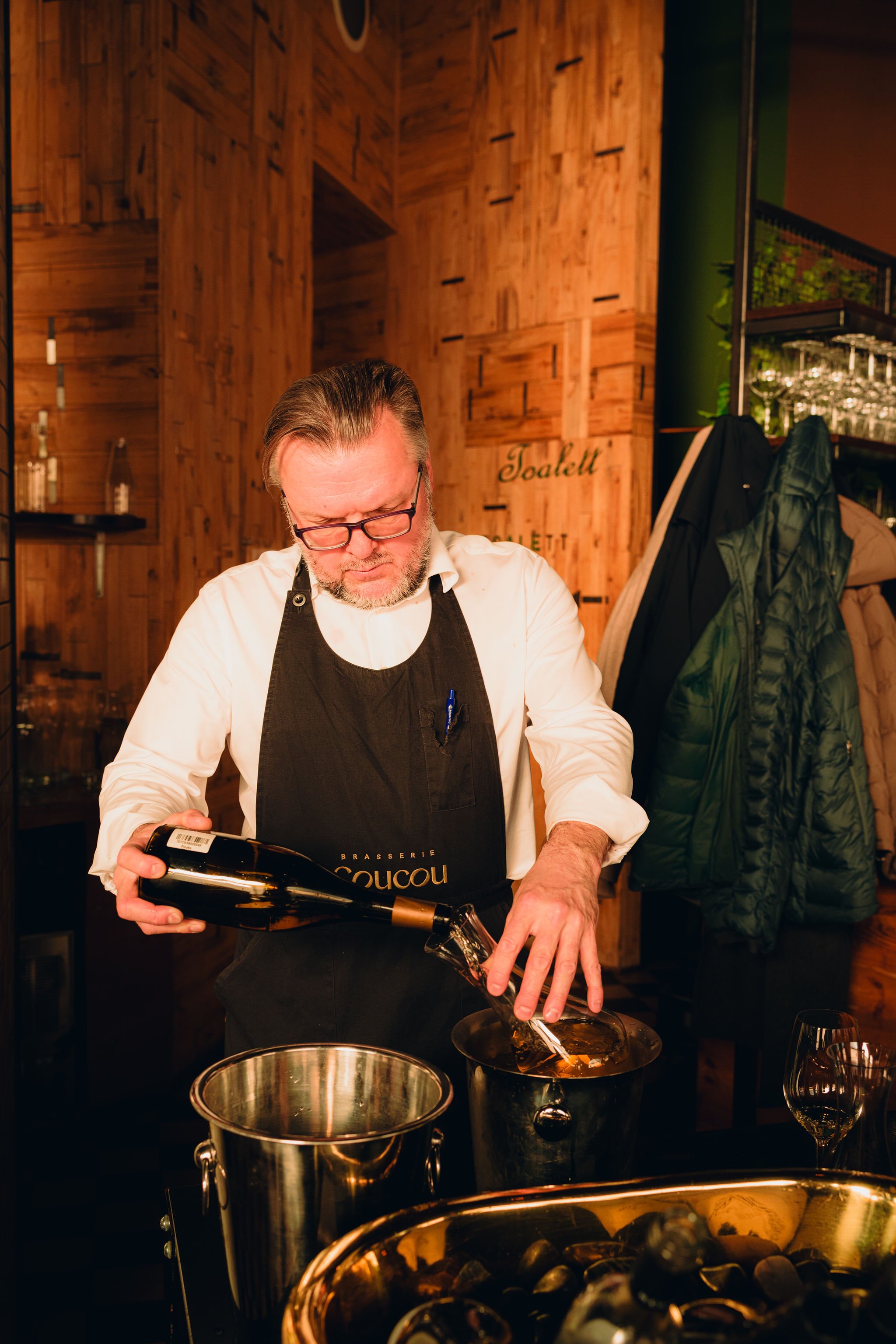 A man with glasses and a beard, wearing a black apron over a white shirt, is pouring wine from a bottle into a glass at a restaurant or wine bar with wooden walls. There are coats hanging on the wall behind him and a table with wine bottles visible in the background.