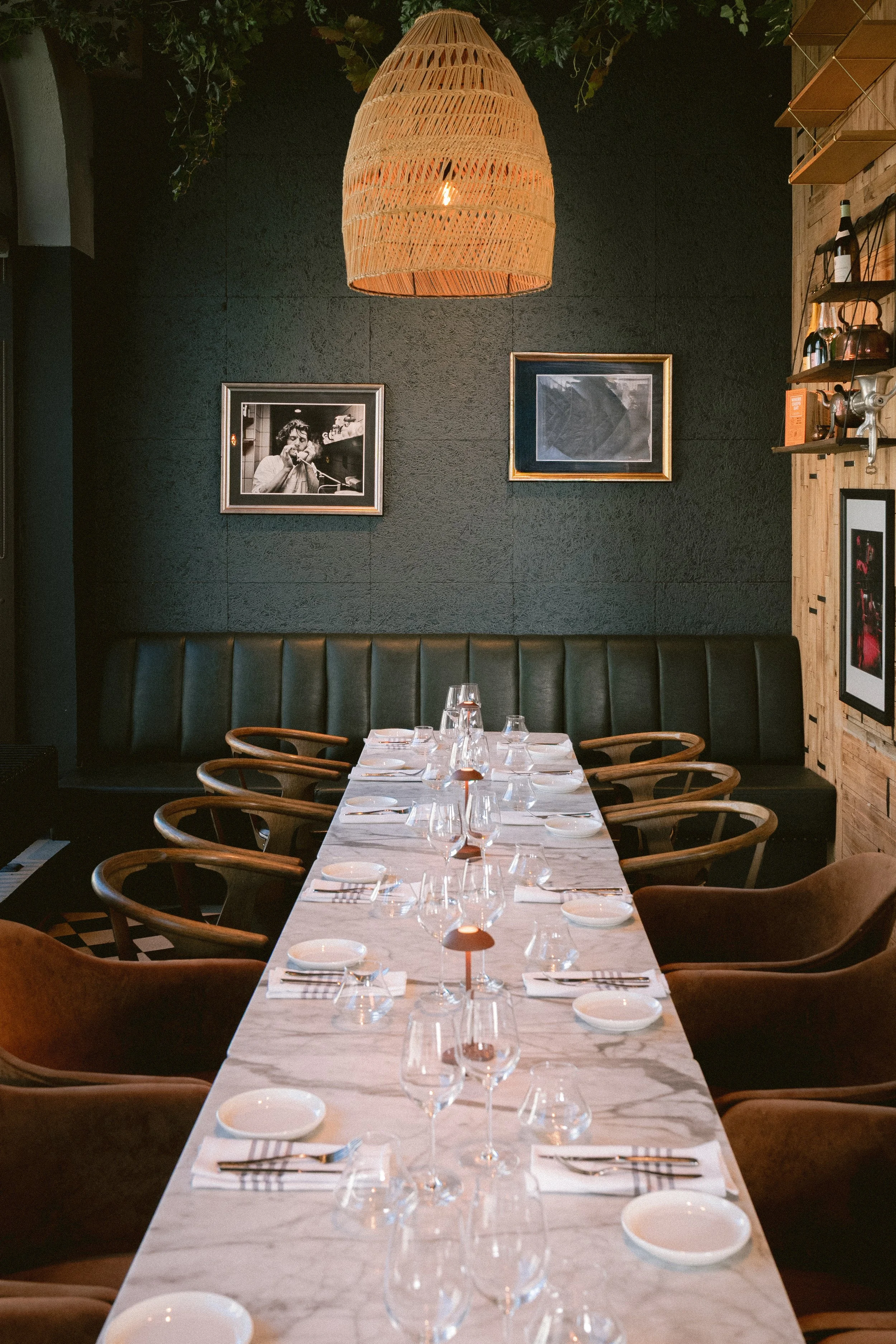 A long dining table set for a meal with white tablecloths, wine glasses, and place settings, located in an upscale restaurant with black and wooden wall decor and framed black and white photos, illuminated by a large wicker pendant light.