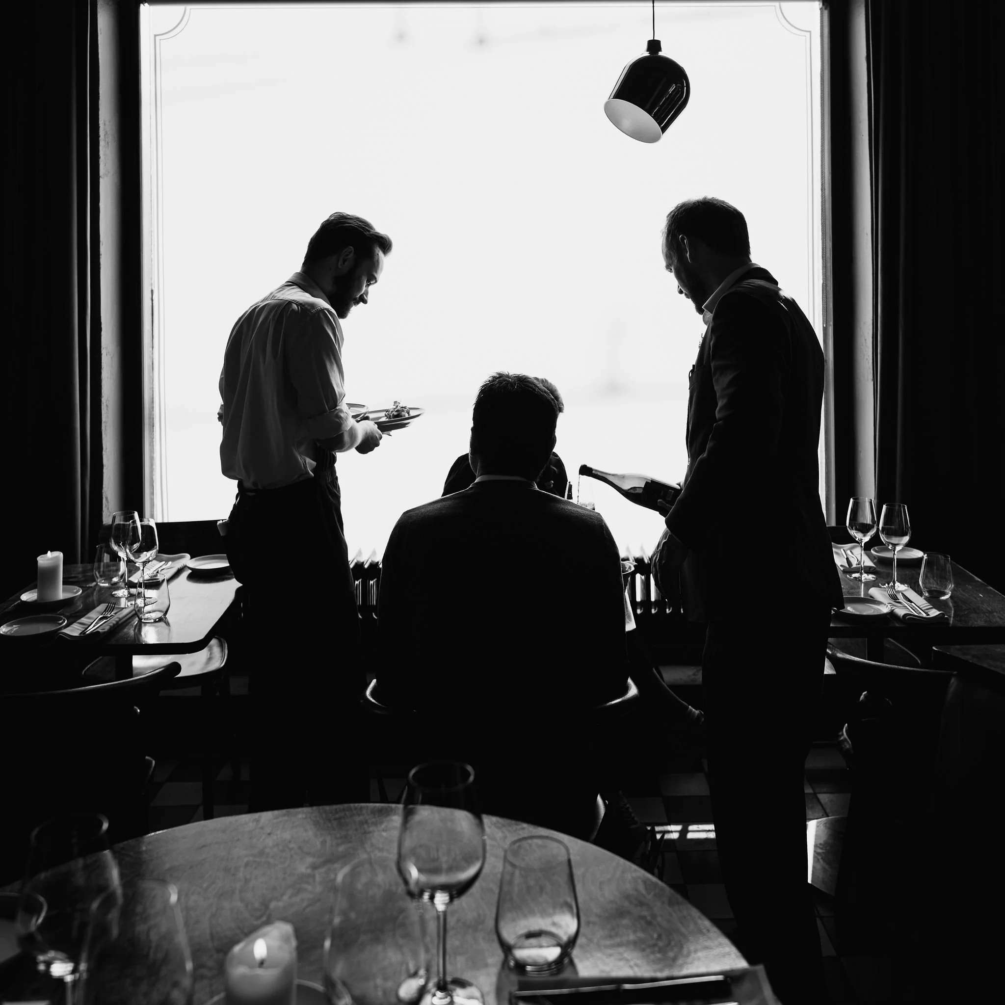 Silhouette of three men in a restaurant, one seated and two standing, near a large window with curtains, dining table set with glasses and silverware.