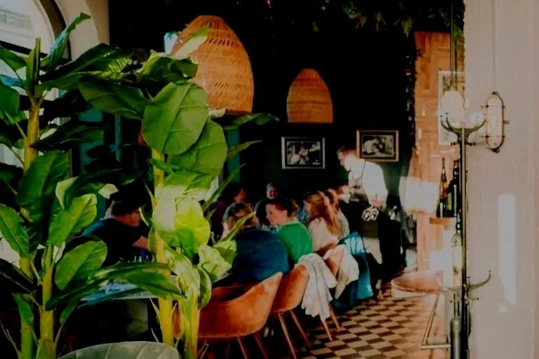 People dining at a restaurant table with large green plants in the foreground, dark walls, framed pictures, and decorative lighting.