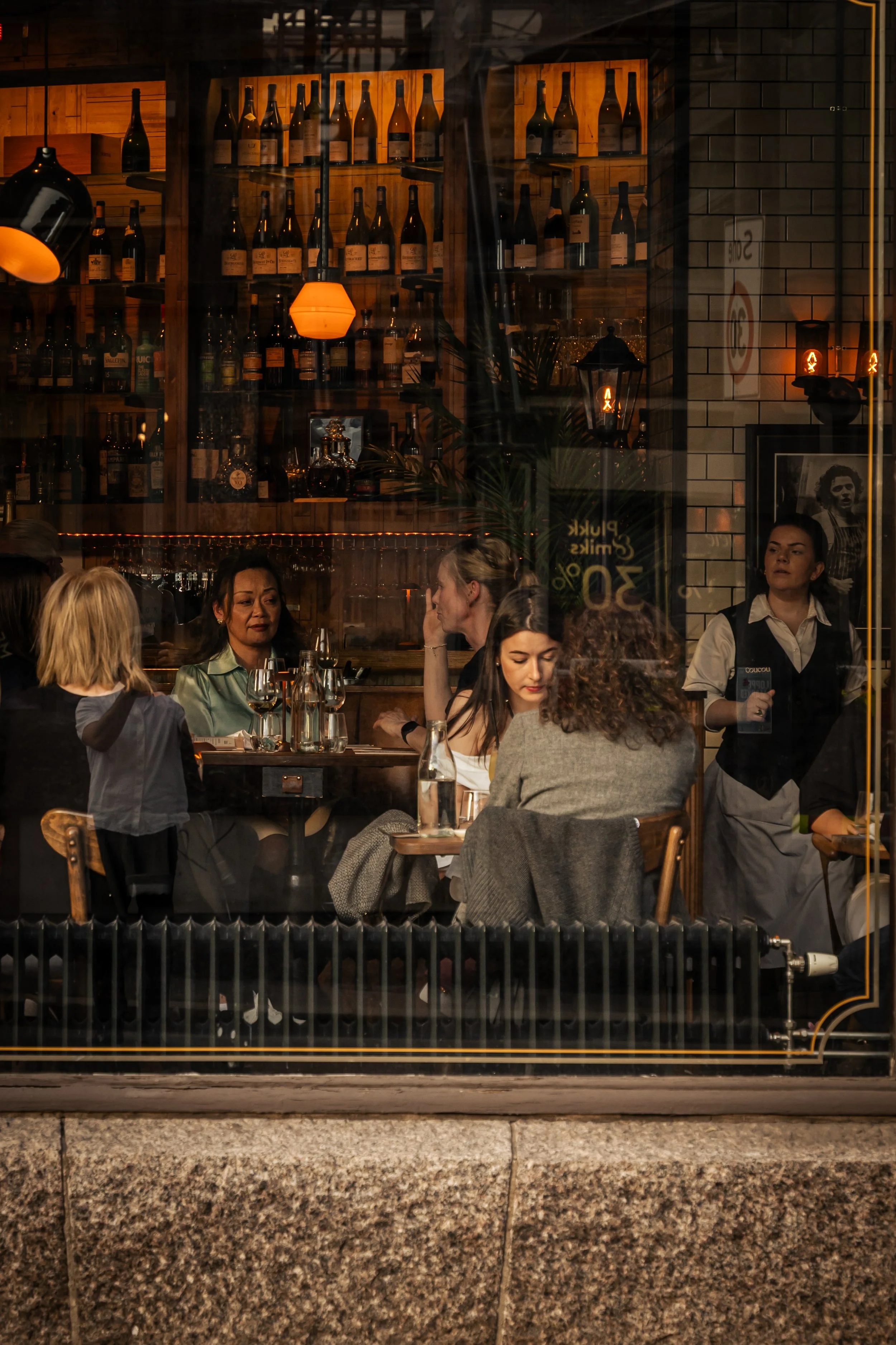 View through a restaurant window showing a group of people dining inside, with a wall of wine bottles on wooden shelves and warm ambient lighting.