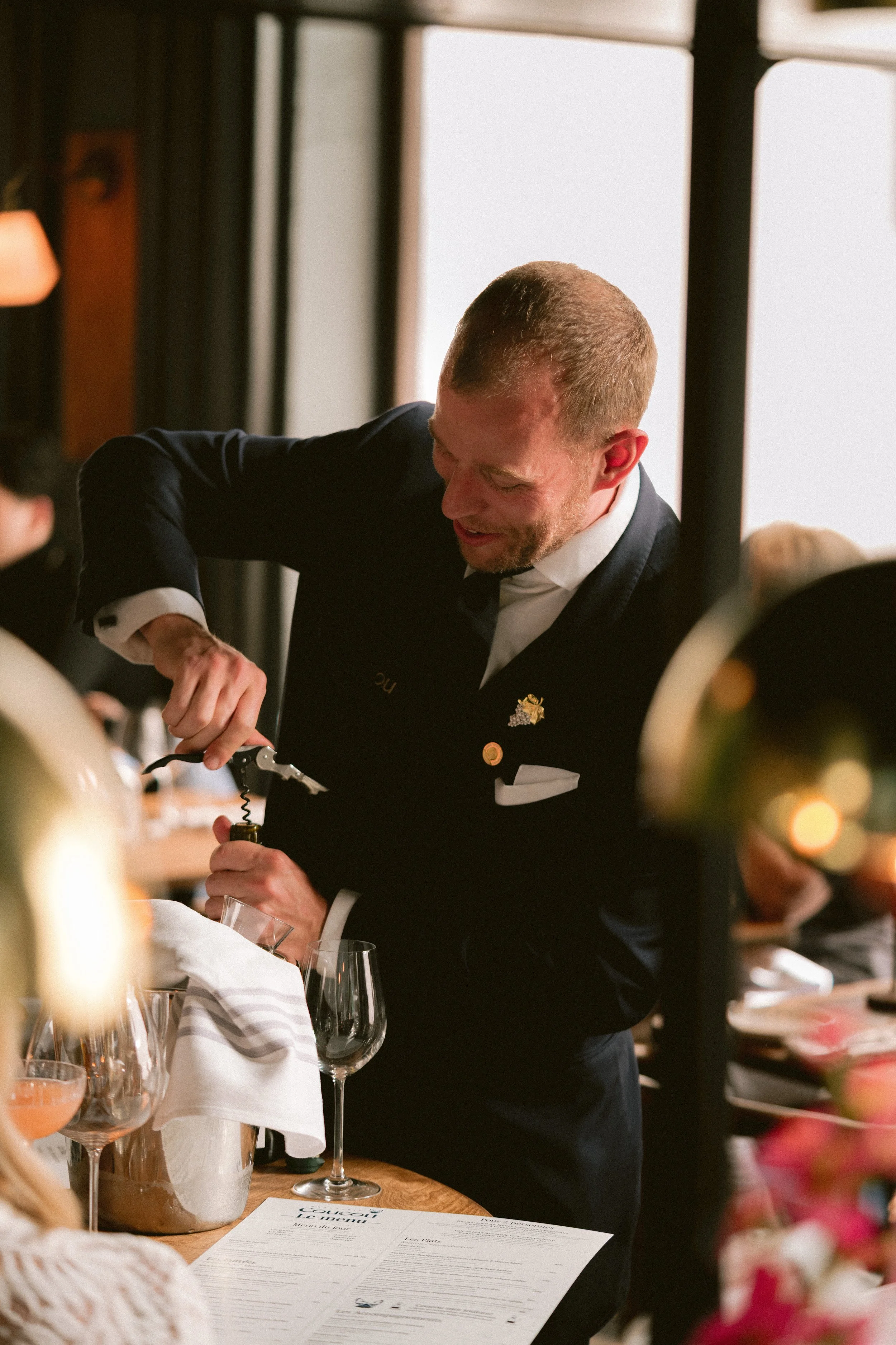 A man in a black tuxedo serving wine at a formal event, with wine glasses, a menu, and floral decorations on the table.