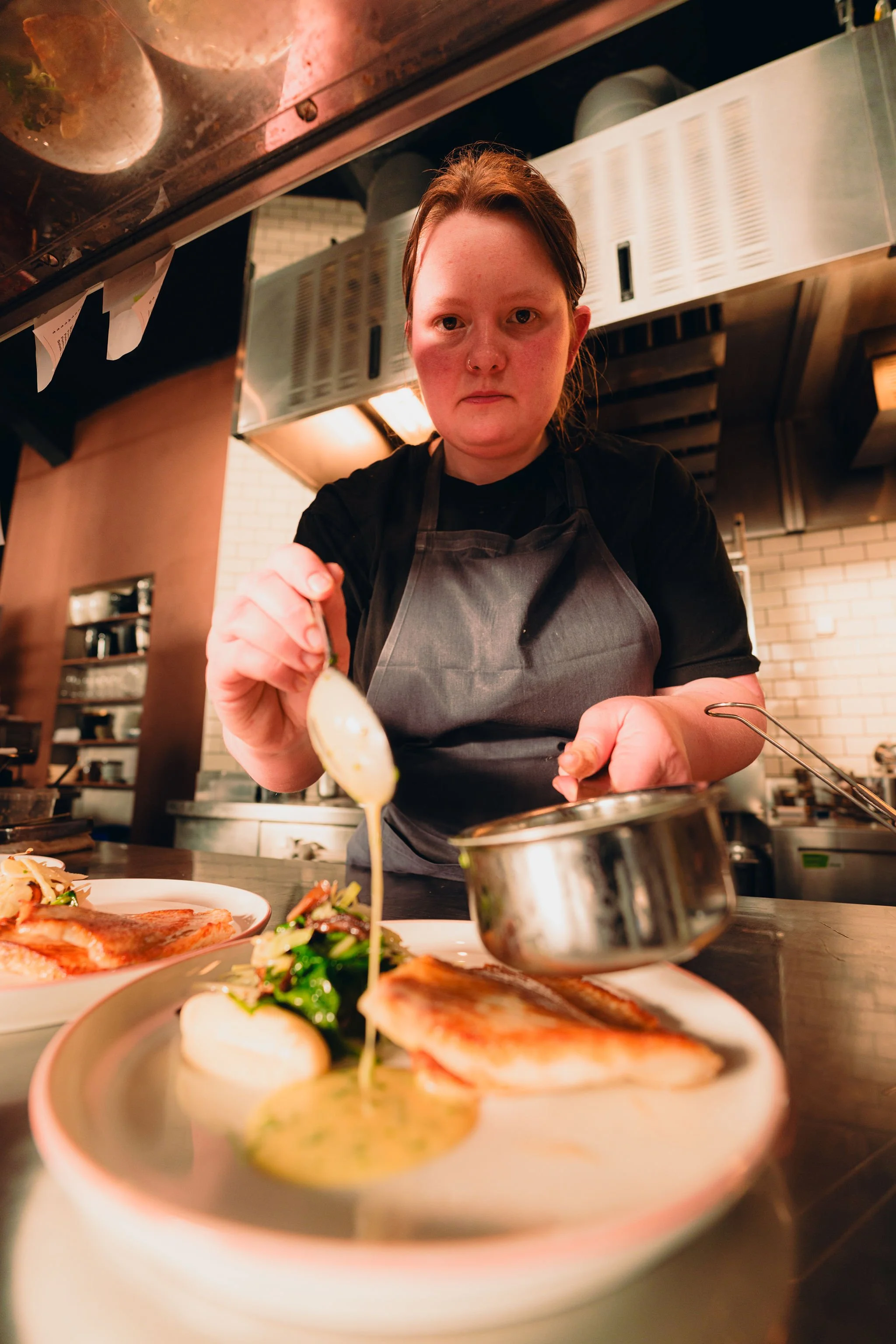 A female chef with red hair and a black apron is plating food in a restaurant kitchen, with plates of prepared dishes including fish, salad, and sauce in the foreground.