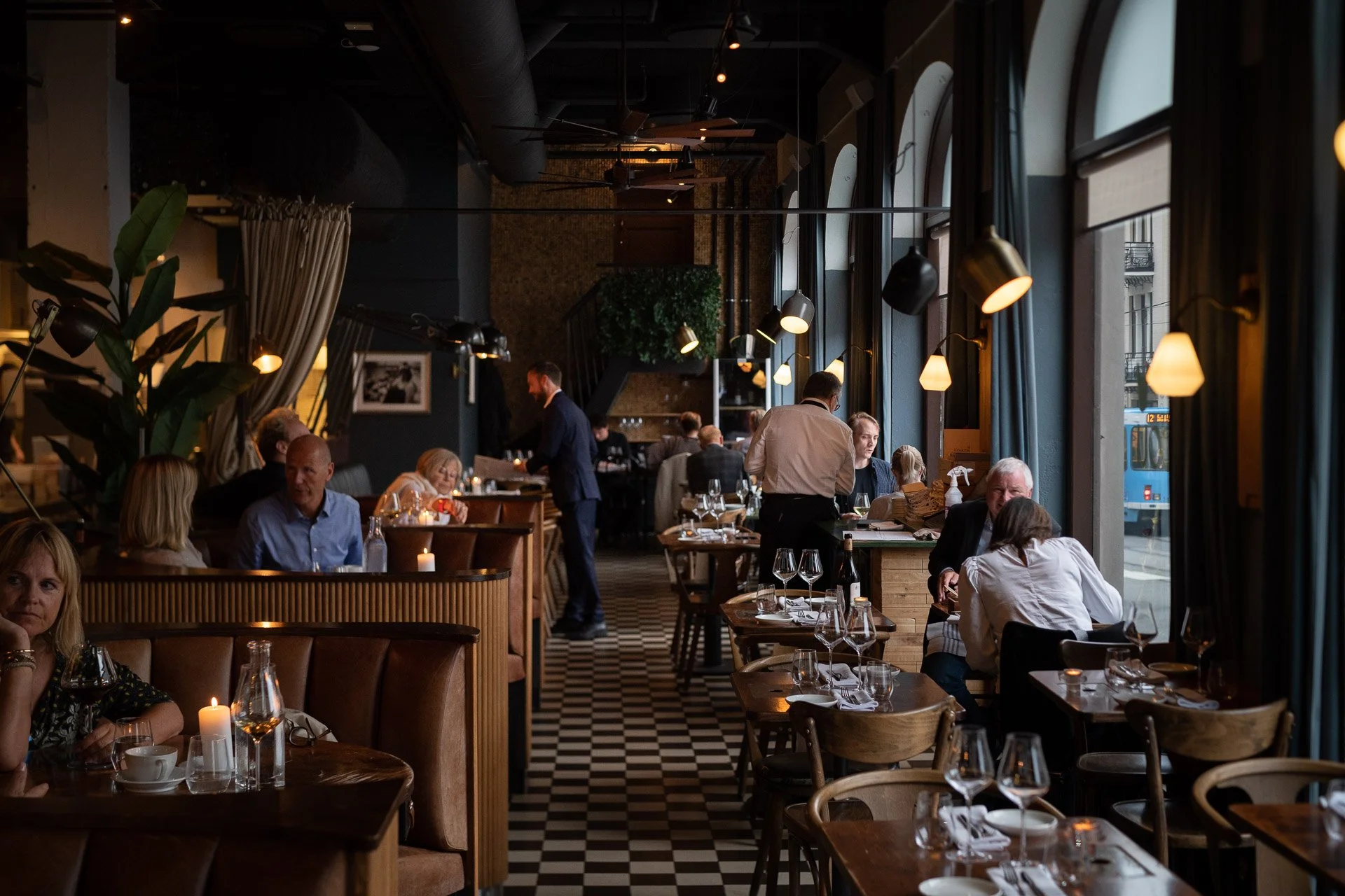 Interior of a restaurant with large windows, hanging lights, and tables set for dining, with customers and waitstaff present.