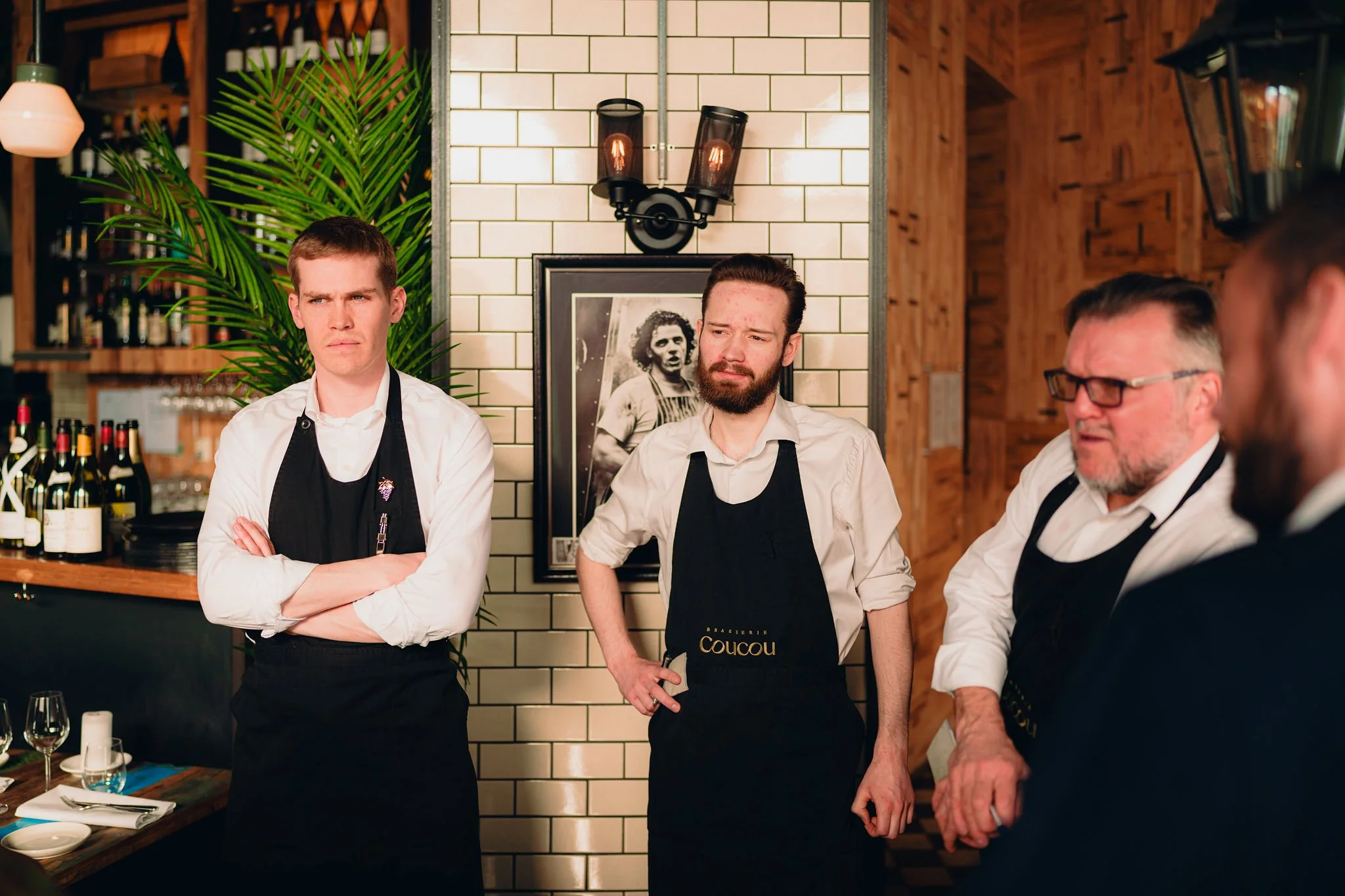Three male restaurant staff members stand in a restaurant setting with wooden and brick decor. Two are wearing white shirts and black aprons, with one crossing his arms and the other with his hands on his hips. The third, partially visible, is wearing glasses. The background features bottles on shelves, a framed black-and-white photo of a woman, a large green plant, and modern light fixtures.