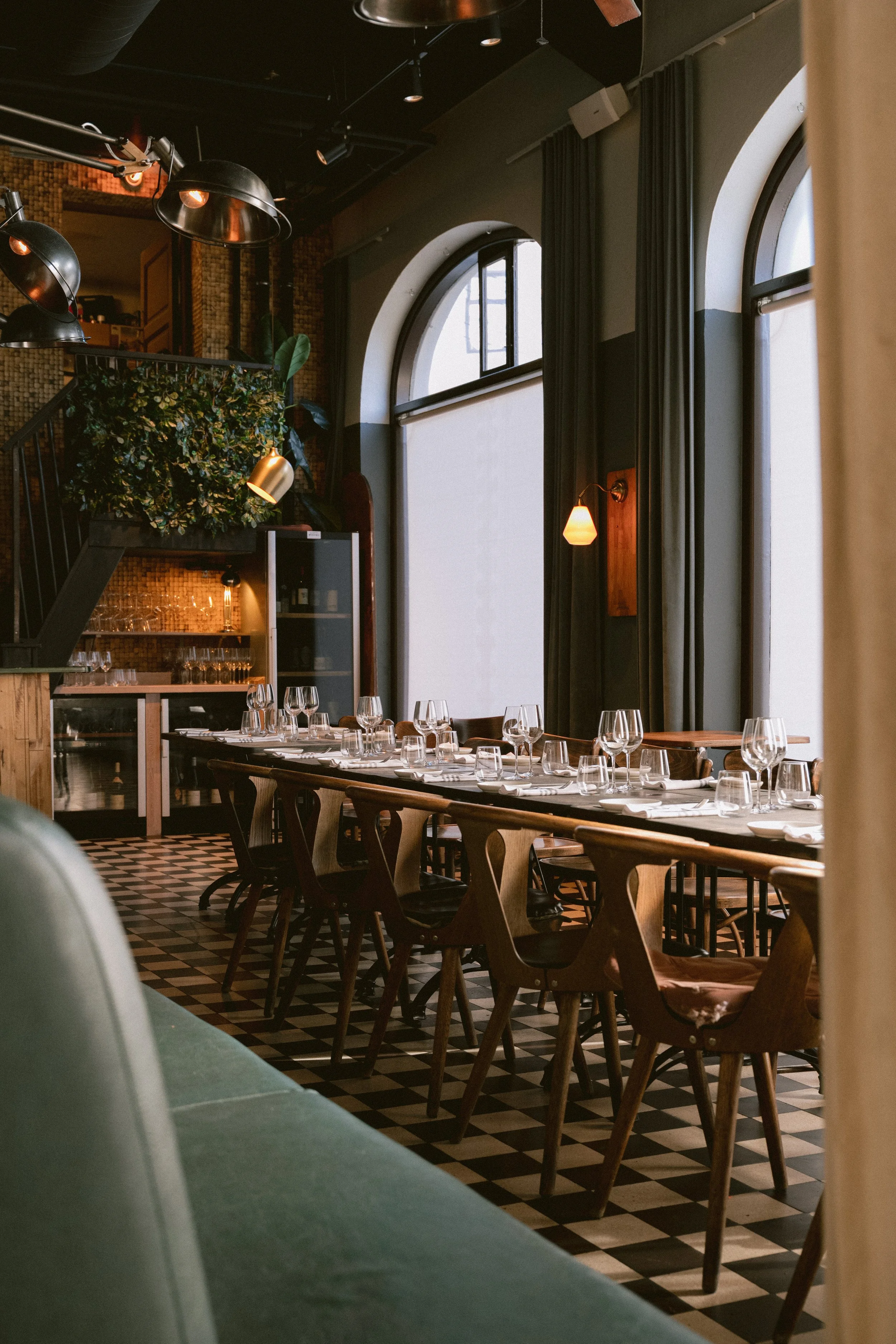 Interior of a restaurant with a long dining table set with glasses and napkins, surrounded by wooden chairs, large arched windows, black curtains, and warm lighting.