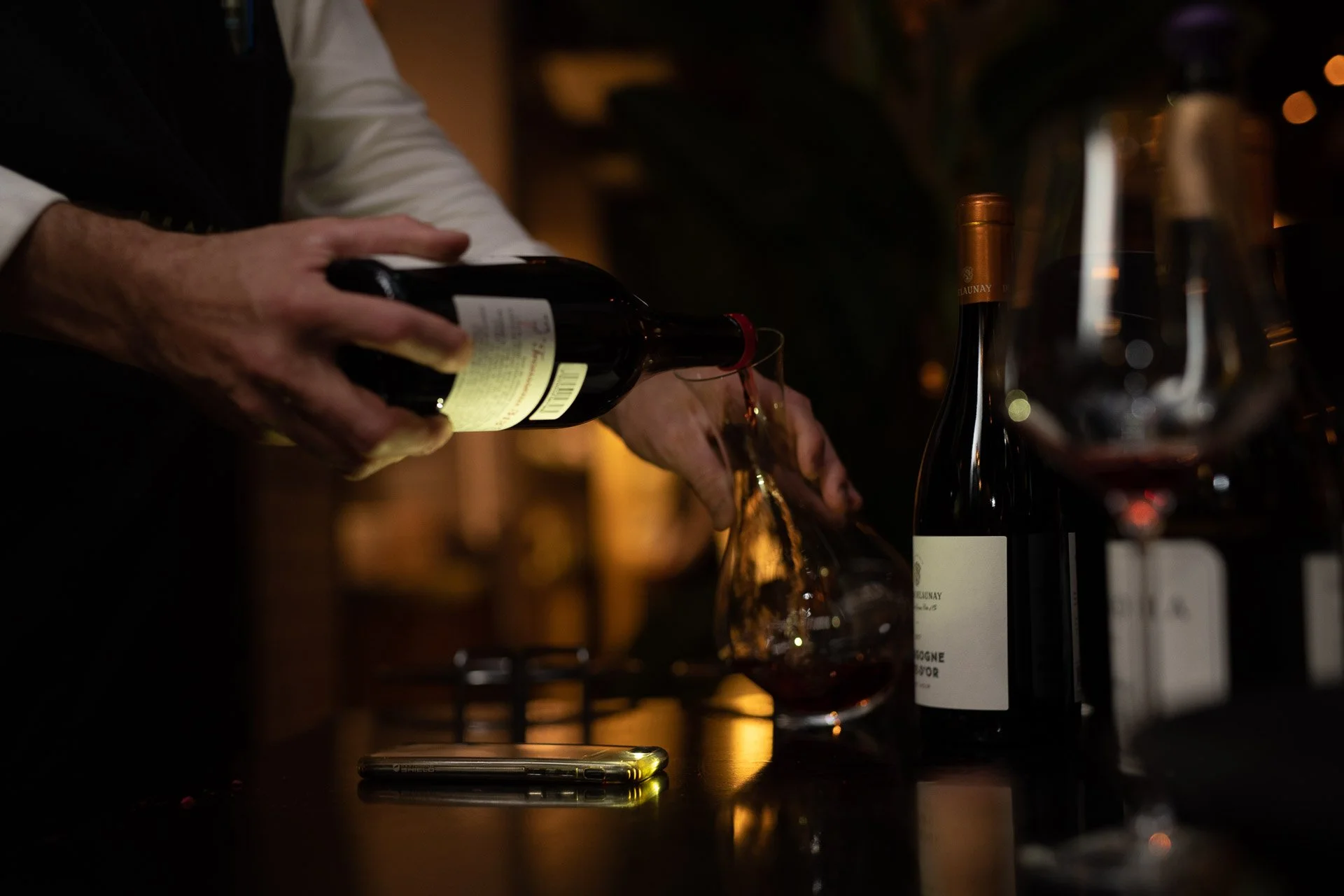 Person pouring red wine from a bottle into a glass in a dimly lit setting, possibly a restaurant or bar.