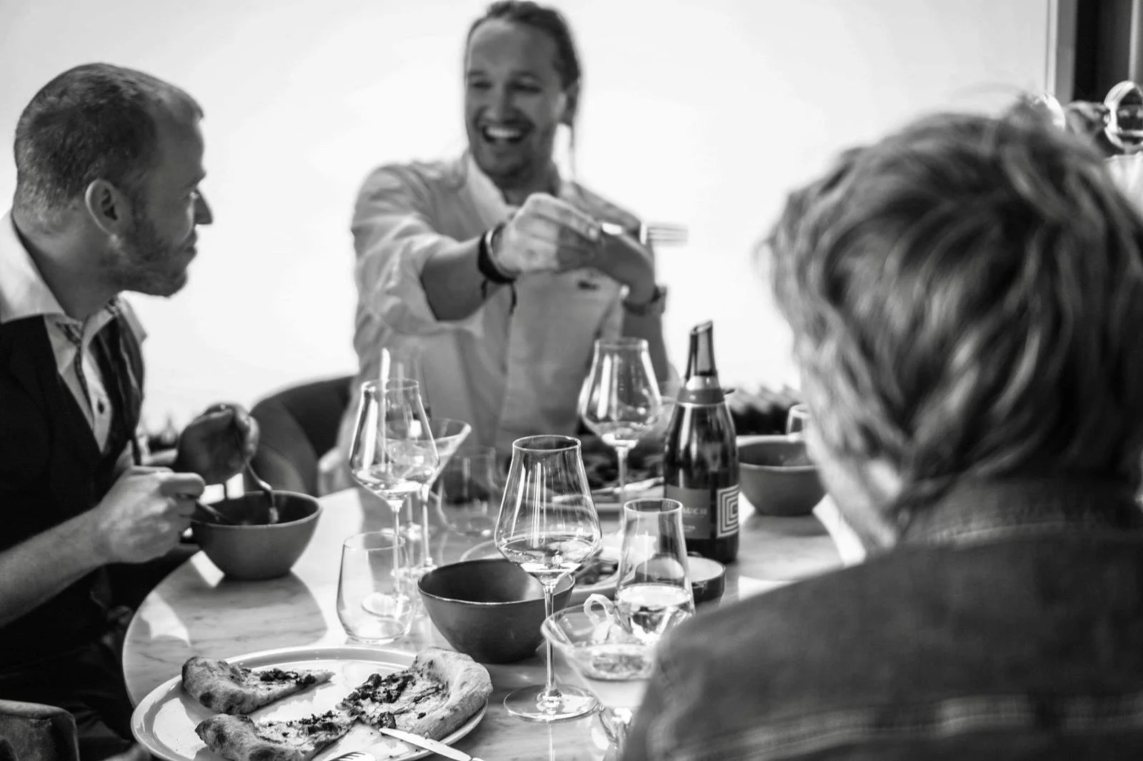 People sitting at a dining table, enjoying a meal and engaging in conversation, with some drinks and pizza on the table.