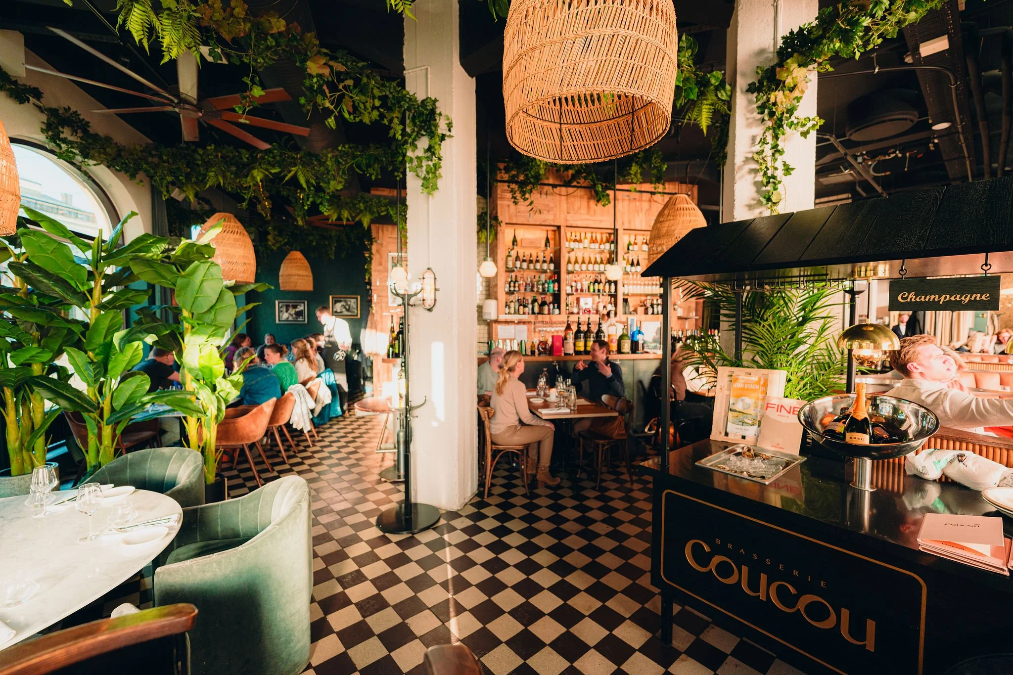 Interior of a lively restaurant with green plants and hanging woven light fixtures, filled with patrons dining and socializing, bar shelves with bottles in the background, and a sign indicating champagne available.