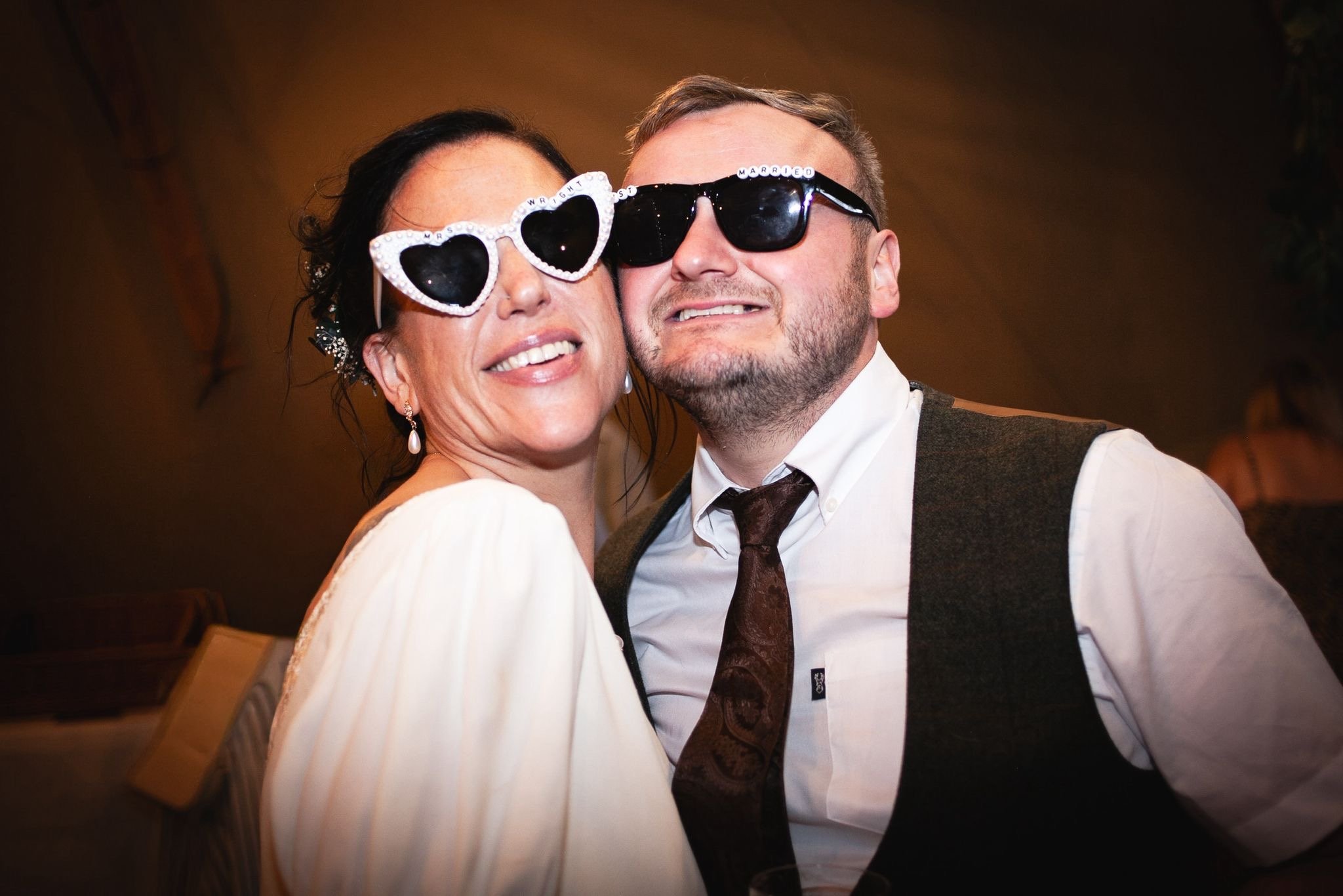 Two people, a woman and a man, wearing large novelty sunglasses, close together and smiling at a celebration or wedding.