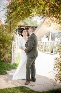 A bride and groom holding hands outdoors near a tree and a wooden fence, with a building in the background.
