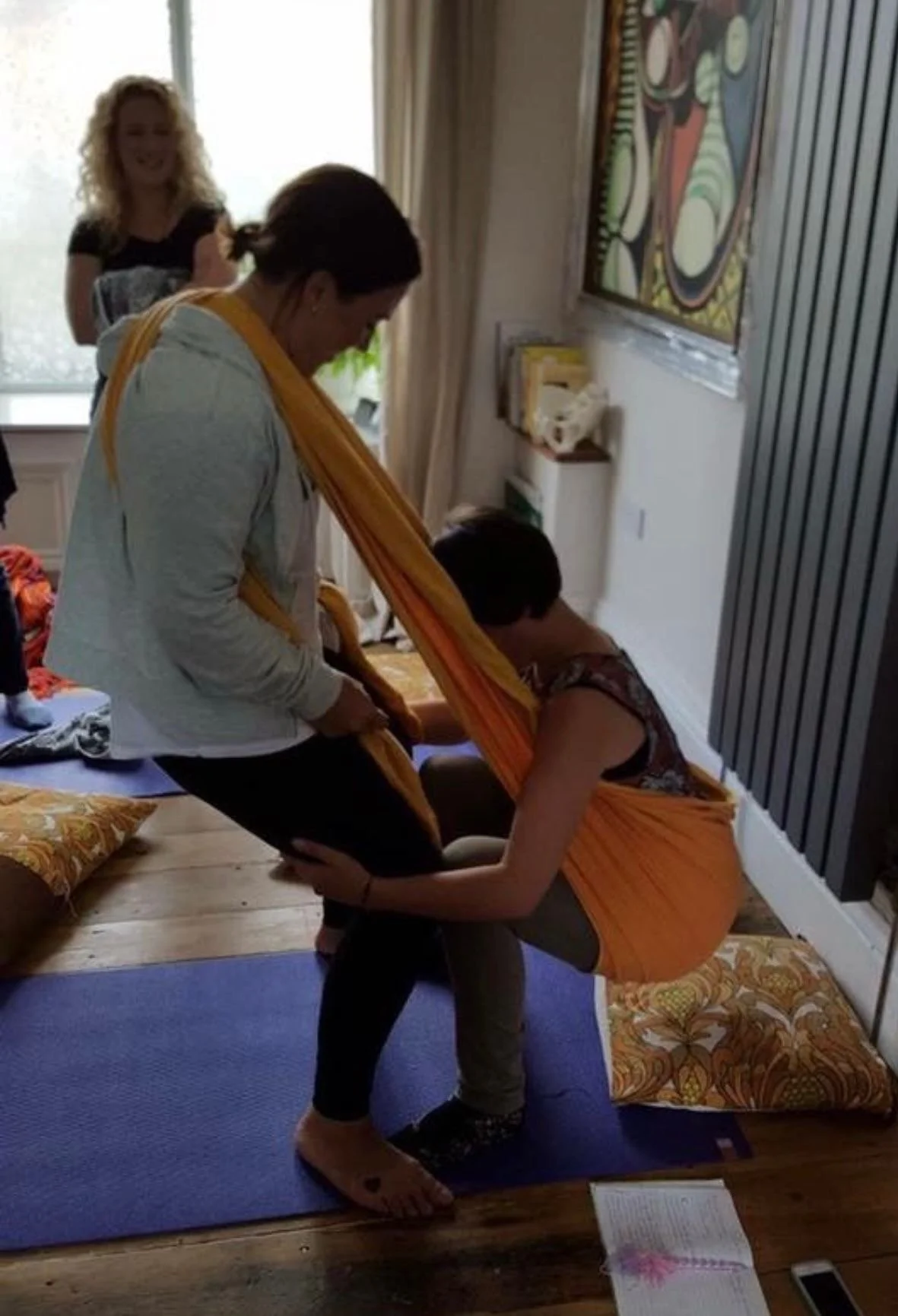 A woman practicing yoga, supported by a trainer, in a home setting with a wooden floor, cushions, and artwork on the walls. There is a trainer in the background smiling.