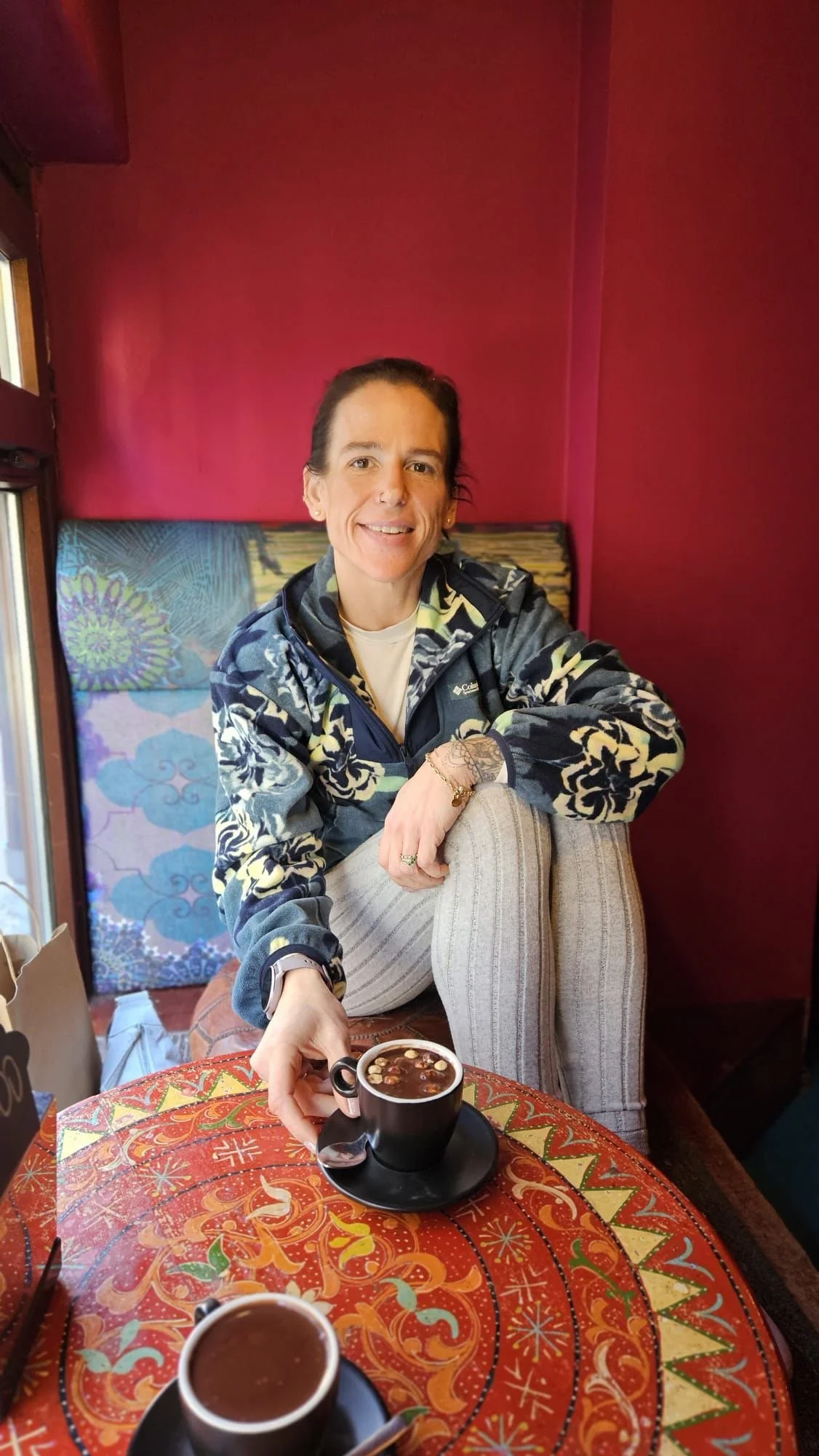 A person with dark hair, wearing a patterned jacket, sitting at a table with a colorful, patterned tablecloth, reaching for a cup of coffee or hot chocolate. The background has a red wall and a quilted headboard or cushion.