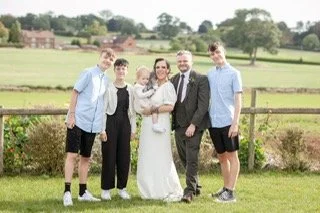 A wedding party of five standing outdoors in a field with a fence and trees in the background. The bride and groom are in the center, with the bride wearing a white dress and holding a bouquet, and the groom in a dark suit. Two young men in light shi