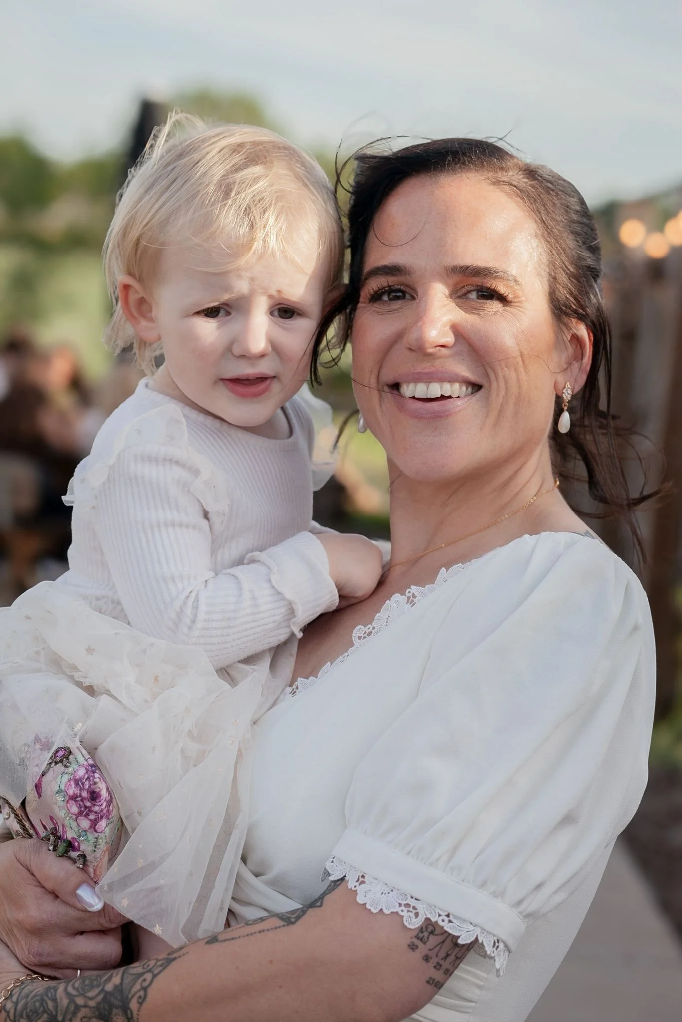 A woman with dark hair, earrings, and a white dress holding a young blonde girl with a worried expression. The background is outdoors with a blurred crowd.