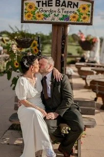 An elderly couple kissing outdoors in front of a garden shop named 'The Barn Garden & Topper', surrounded by flowers and outdoor furniture.