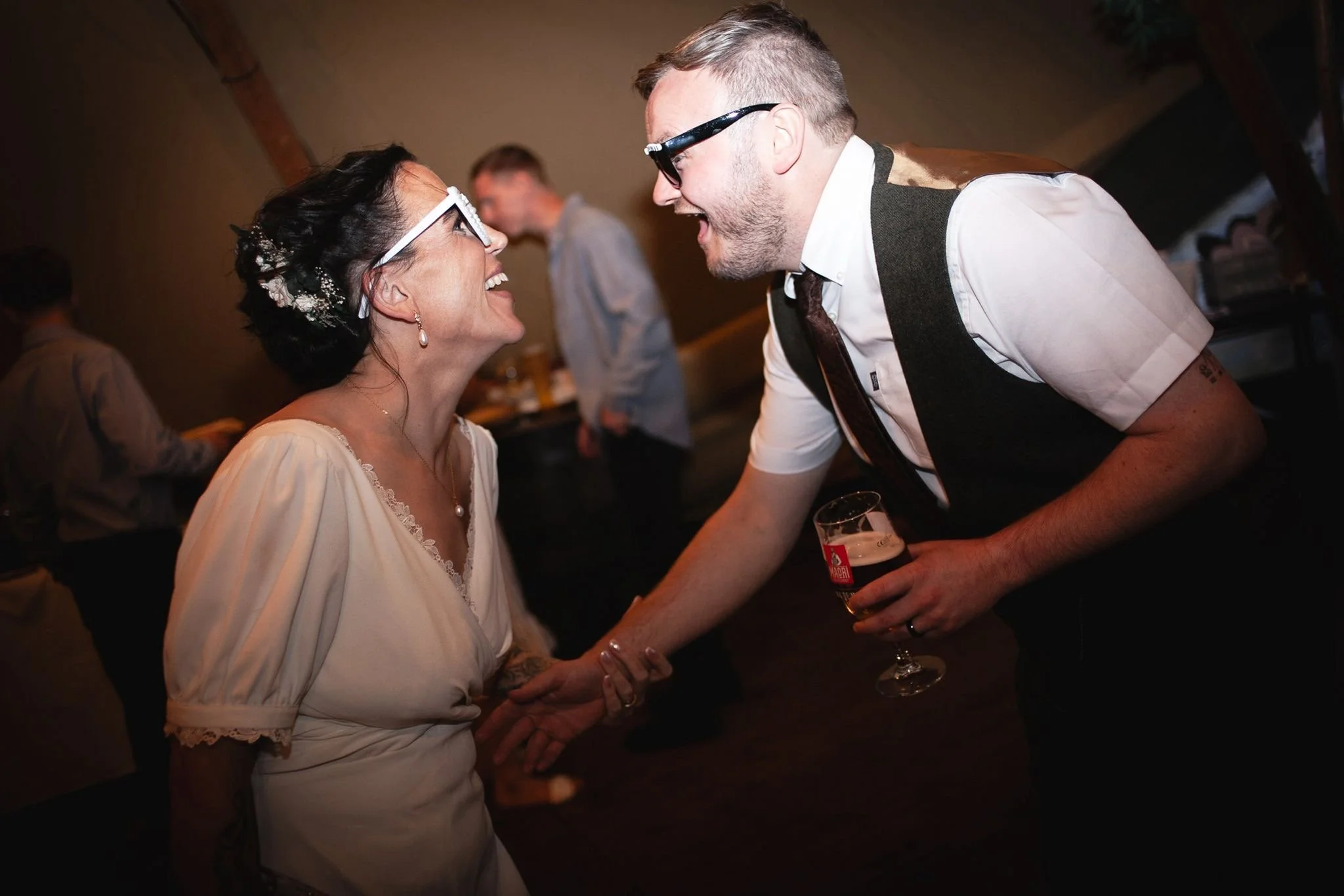 A woman and a man are smiling and holding hands at a celebration, possibly a wedding or party, with other guests in the background.