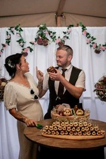 A couple celebrating a wedding, standing in front of a decorated backdrop with cake and desserts on a table.