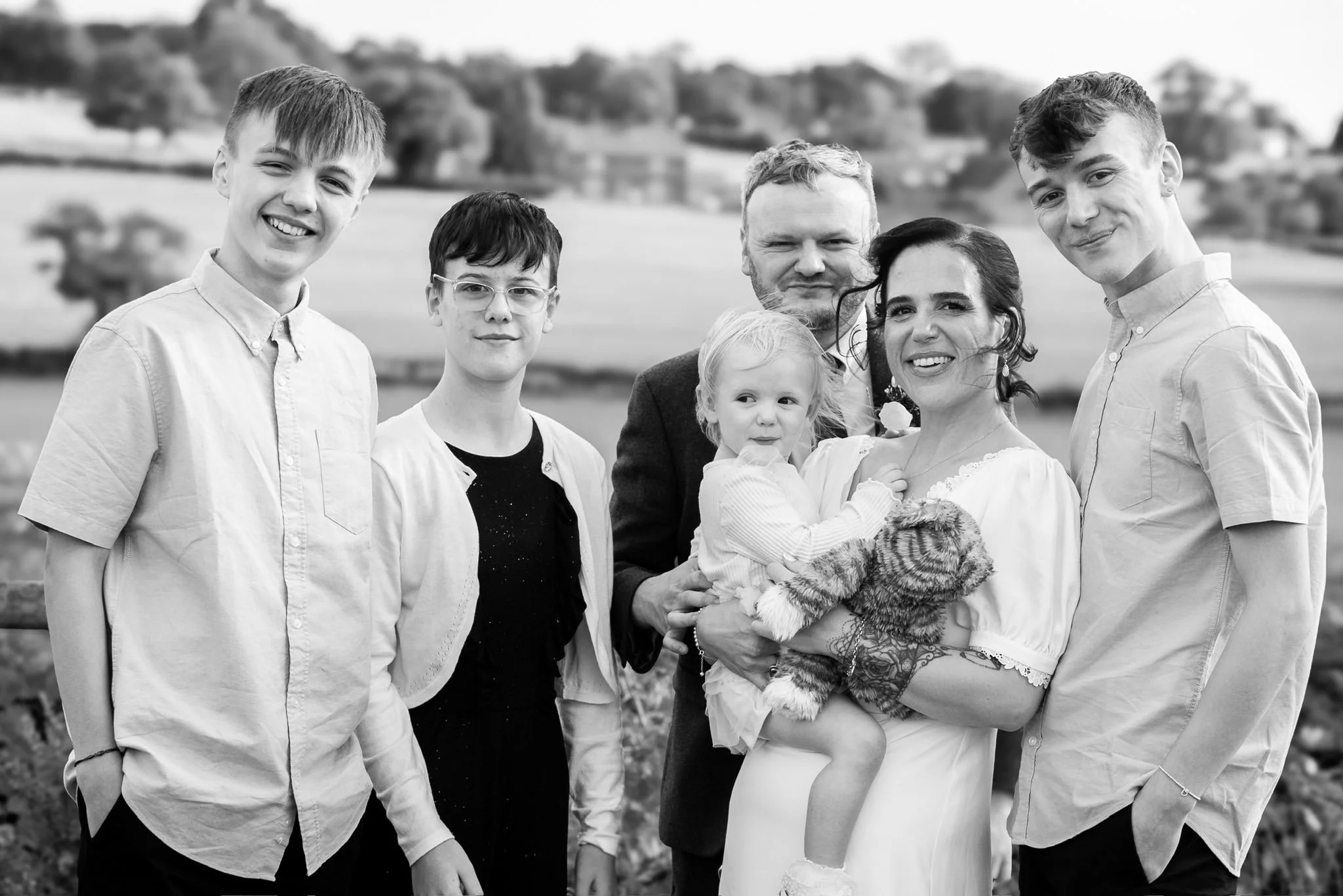 A black and white photo of a large family group outdoors, including adults and children, smiling and posing together in a scenic rural setting with fields and trees in the background.