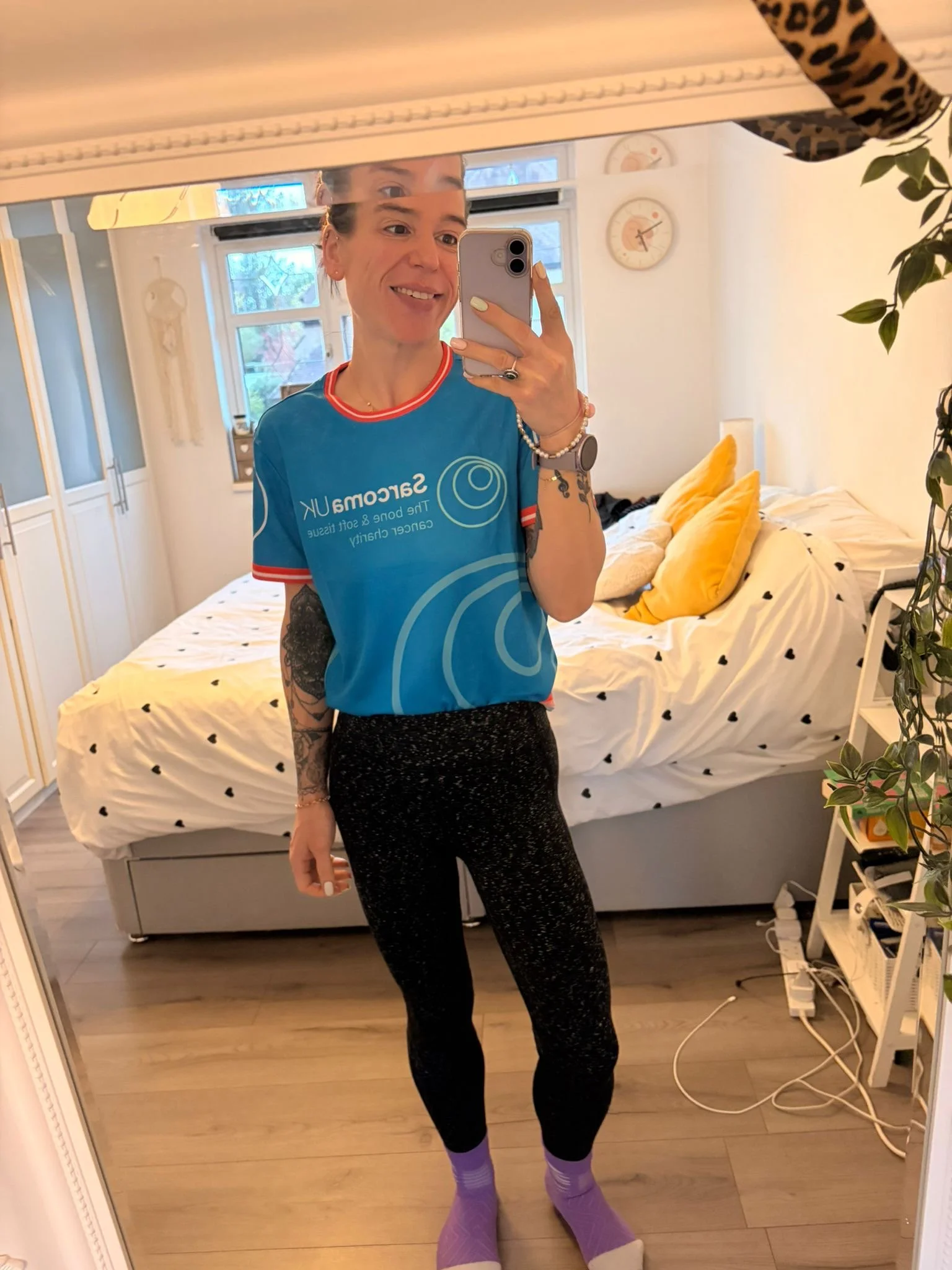 A person taking a mirror selfie in a bedroom, smiling, wearing a blue sports jersey, black sparkly pants, and purple socks, with a bed, pillows, and window in the background.