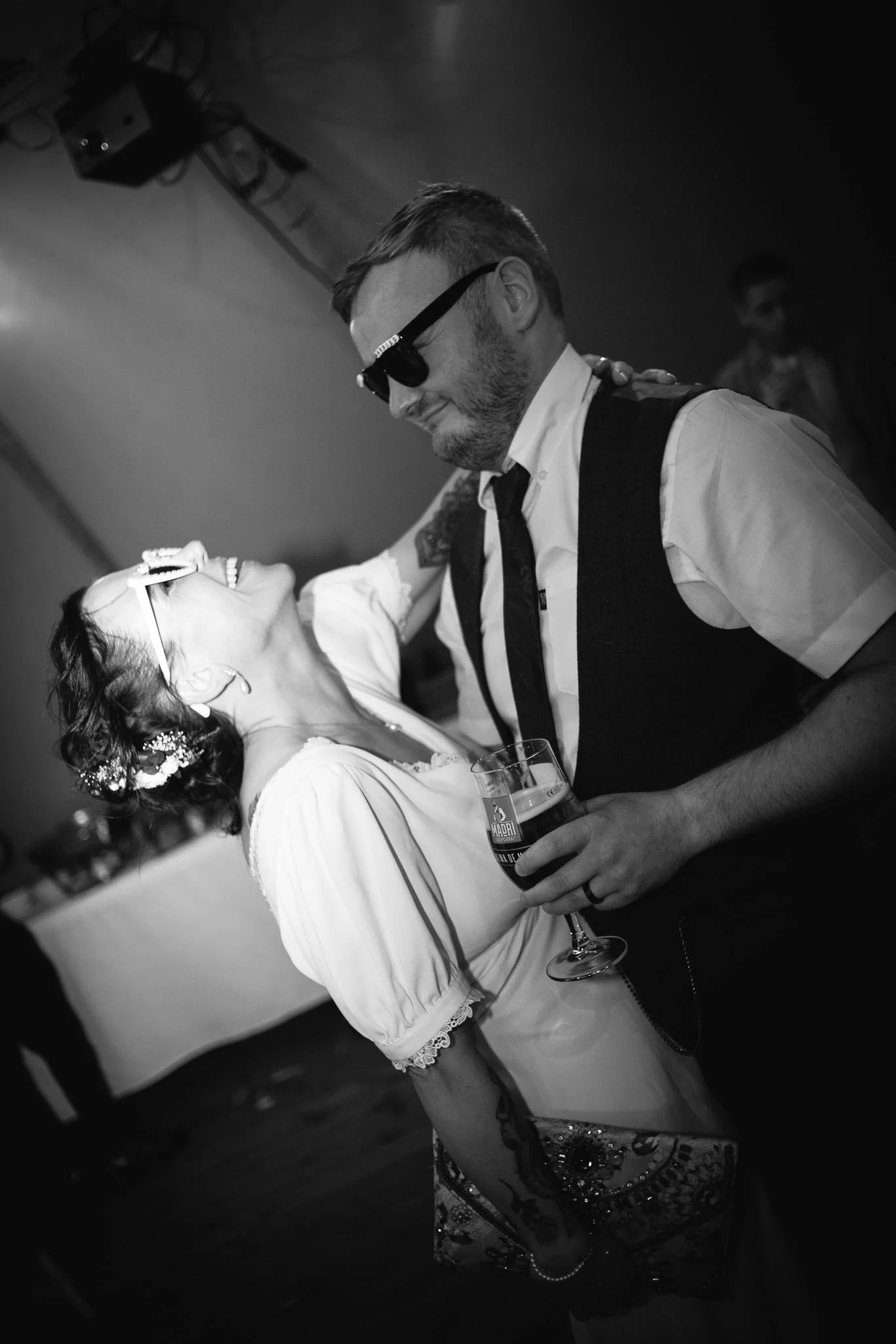 A man and woman are dancing, smiling, and looking at each other at a celebration or party. The woman has floral hair accessories and is holding a glass of beer, wearing a white blouse with lace sleeves. The man is wearing sunglasses, a white shirt, a