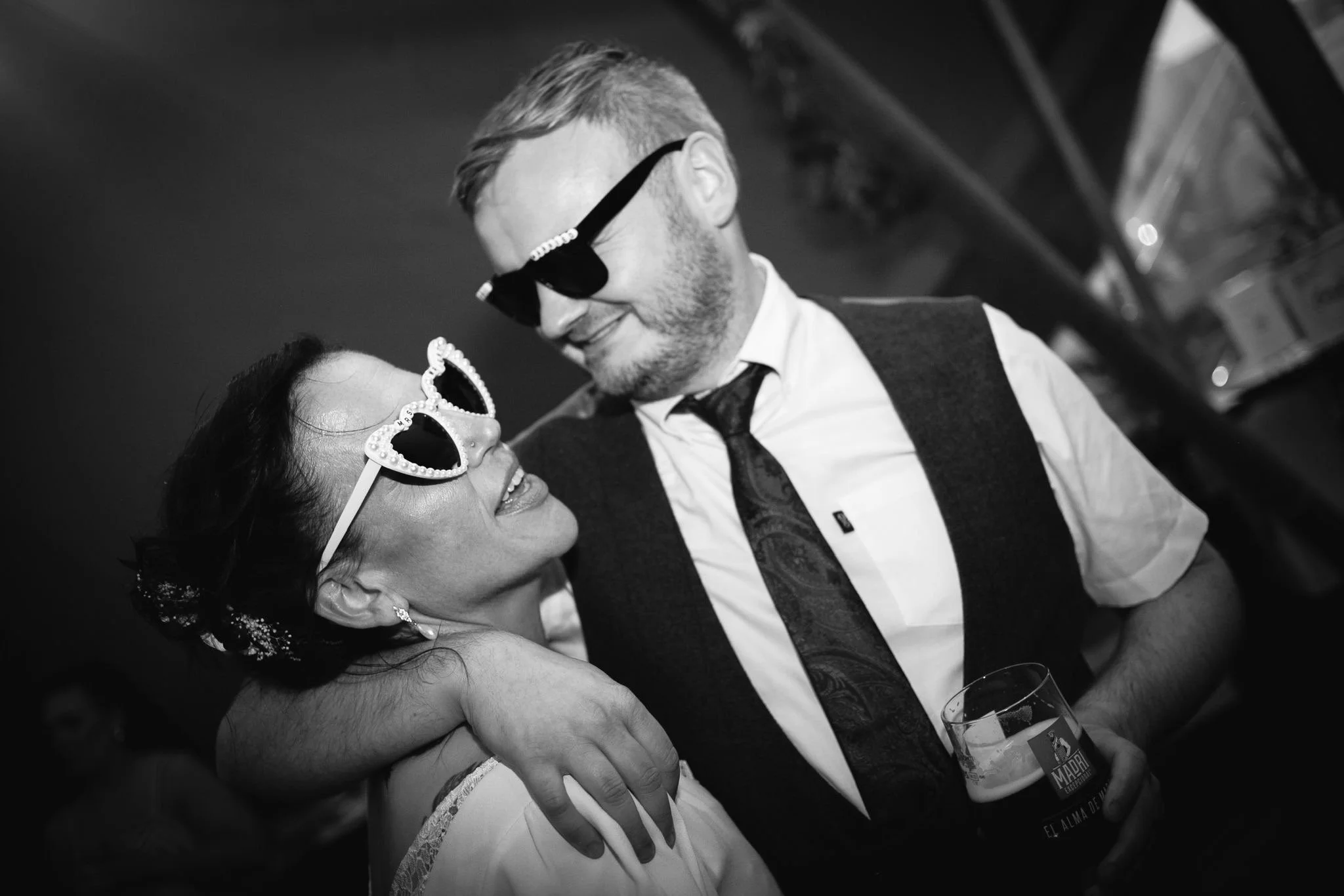 A man and a woman are smiling and looking at each other while dancing together at a celebration. The woman is wearing sunglasses with a heart-shaped frame and a dress, and the man is wearing a shirt, tie, vest, and sunglasses. They are holding hands 