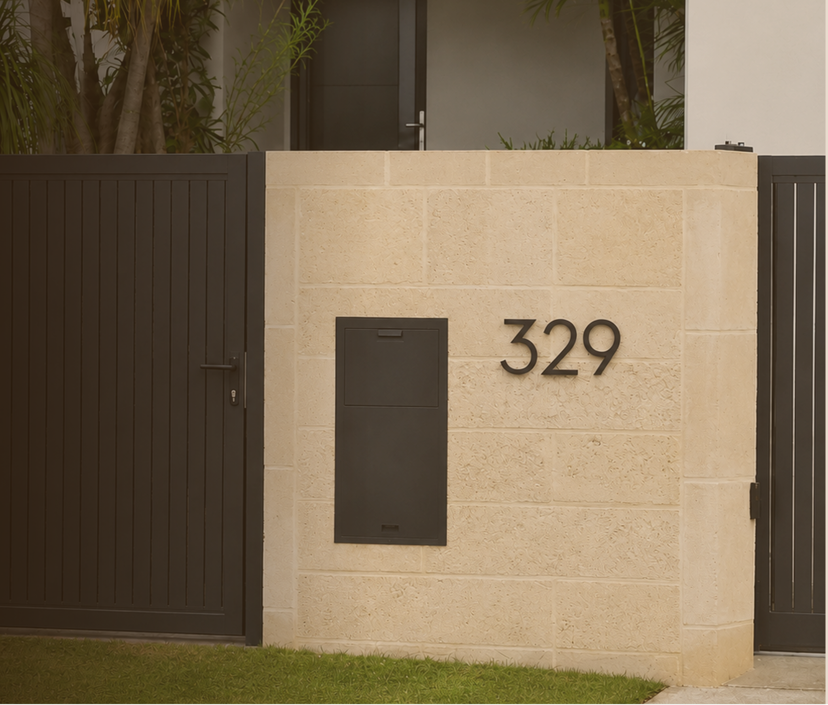 Photo of a modern house entrance with a limestone wall displaying the house number 329 in anthracite numbers, an anthracite mailbox, a monument steel gate on the left, and lush greenery in the background.