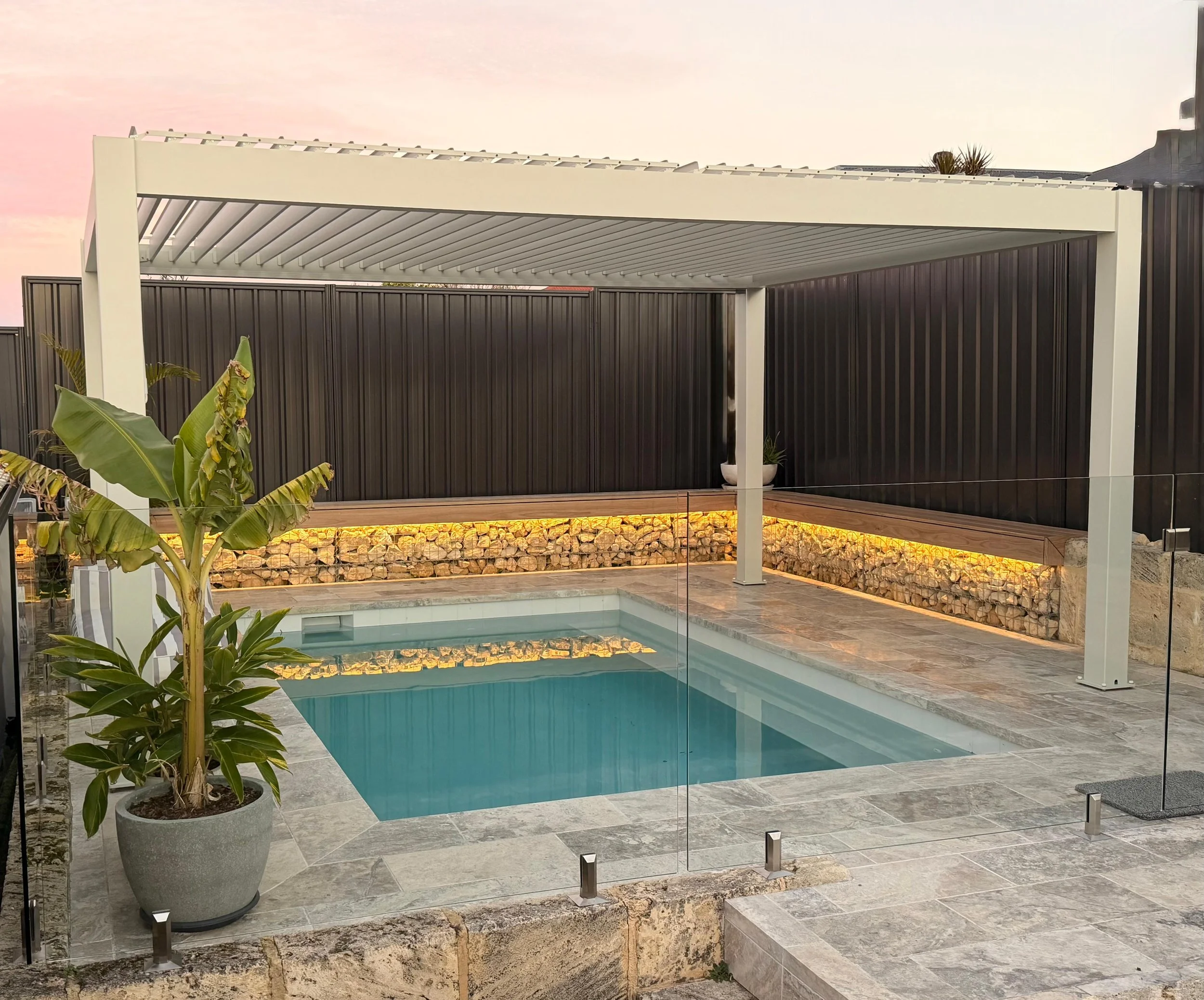 Modern backyard with a swimming pool, potted tropical plant, and a bioclimatic pergola in white, surrounded by a monument boundary fence and decorative gabion wall.
