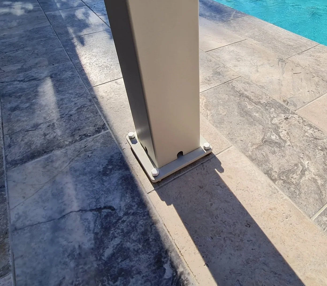 Close-up of a metal support post on a tiled poolside area, casting a shadow on the tiles with part of a swimming pool visible in the background.