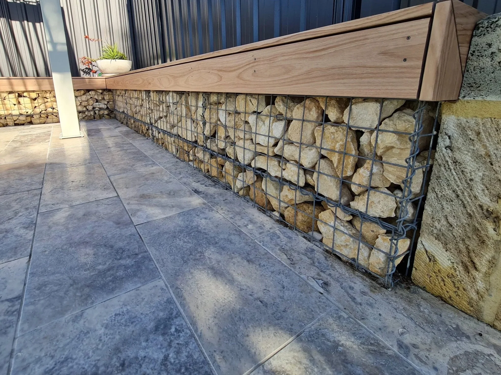 A wooden bench with a simple rectangular design, set on a stone patio, supported by a gabion filled with large rocks, situated against a Monument metal fence. A small ceramic pot with some plants is on the bench.