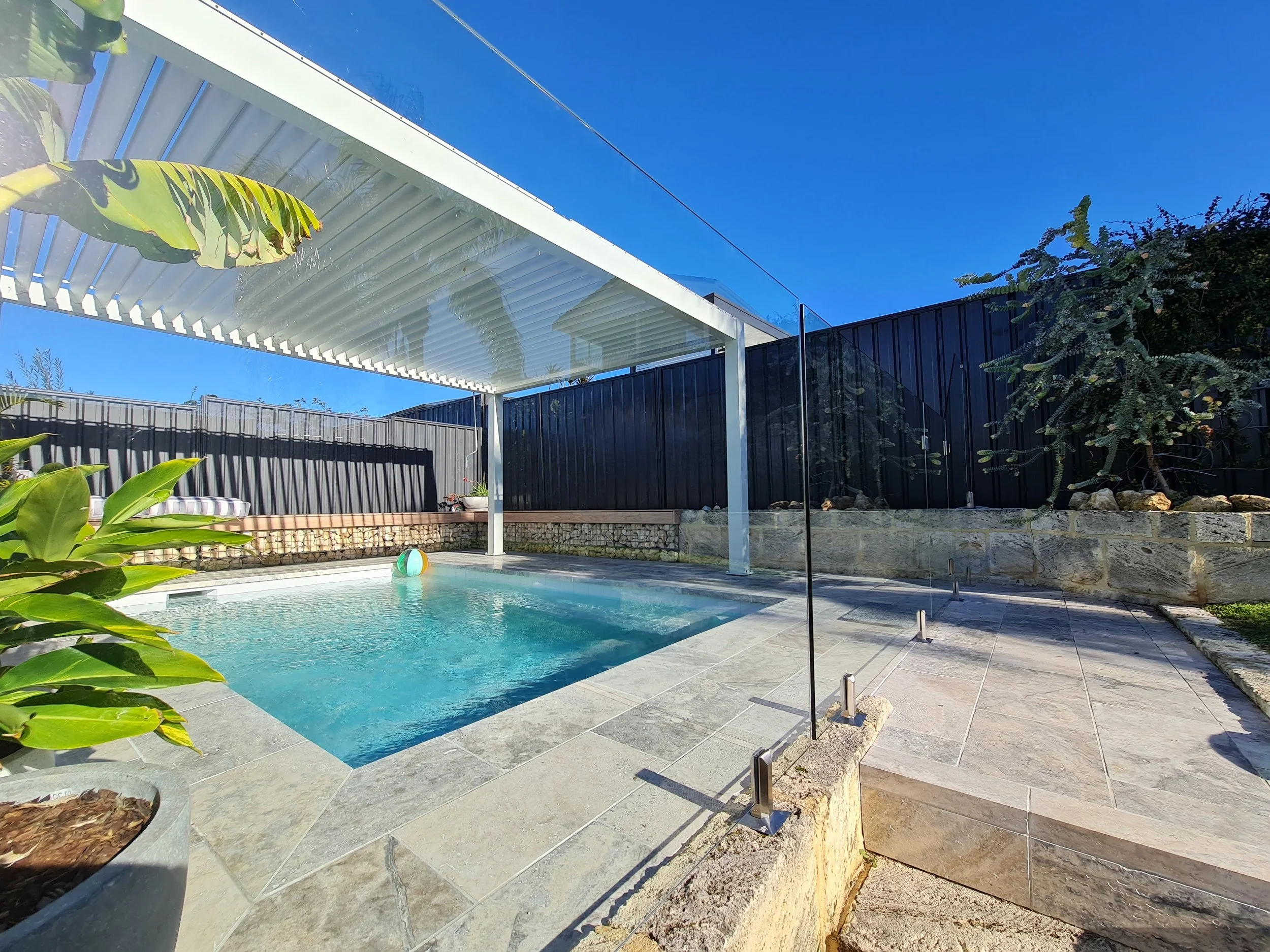 A backyard swimming pool with a modern house and blue sky in the background. The pool has a transparent safety fence, greenery, and a shaded patio area.