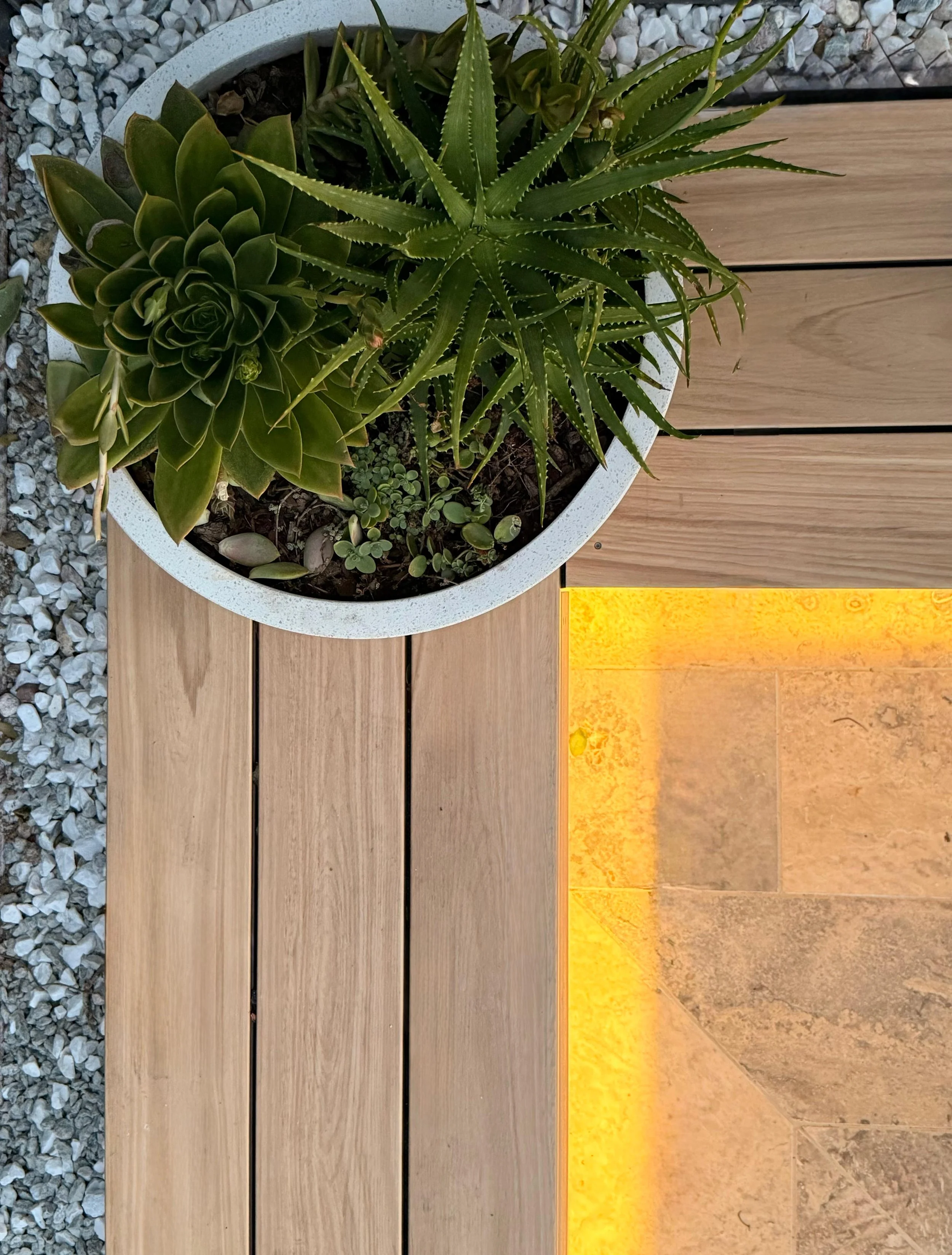 An overhead view of potted succulents on a composite decking bench seat, with travertine paving lit up by LED strip lighting