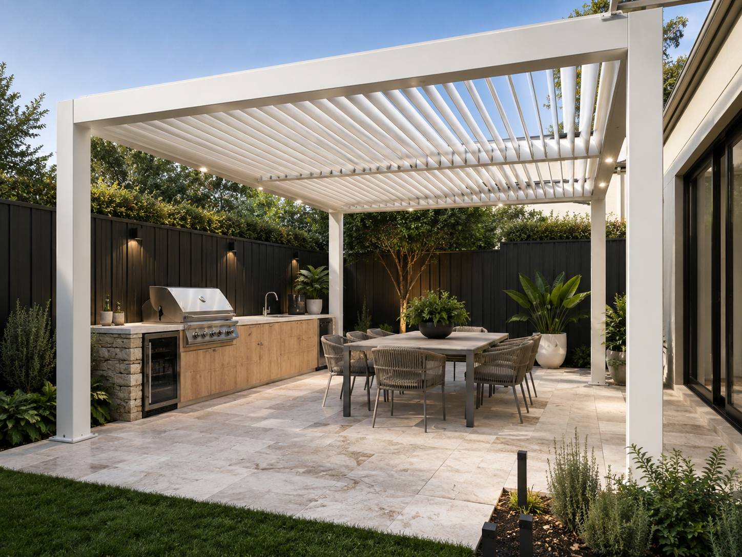 Modern outdoor patio with a white pergola, dining table with chairs, outdoor kitchen with grill, potted plants, and garden against a black fence and trees.