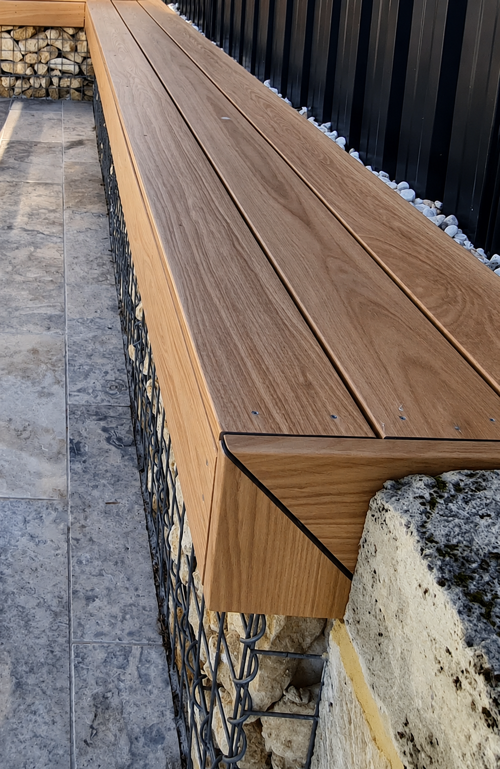 A composite decking beachseat, with limestone gabion wall and silver travertine paving in background 