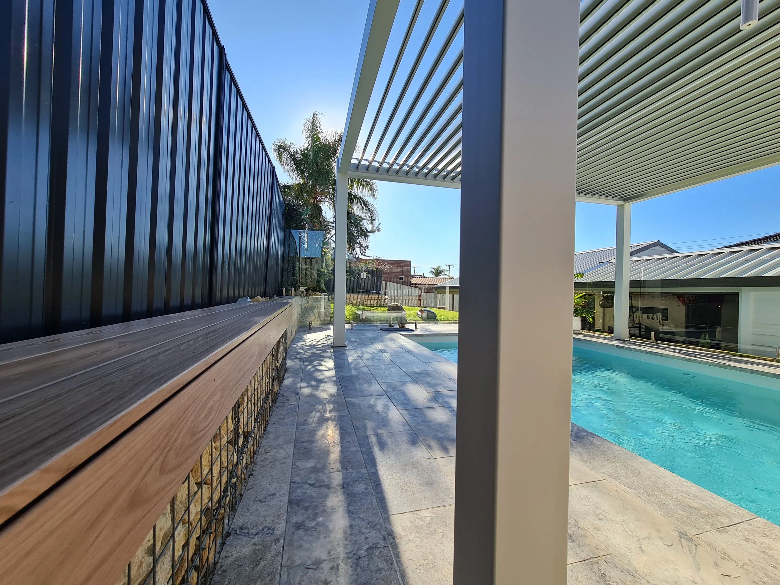 View of a backyard with a swimming pool, modern architectural elements, a black metal fence, and a patio with gray tiles, palm trees, and neighboring houses in the background.