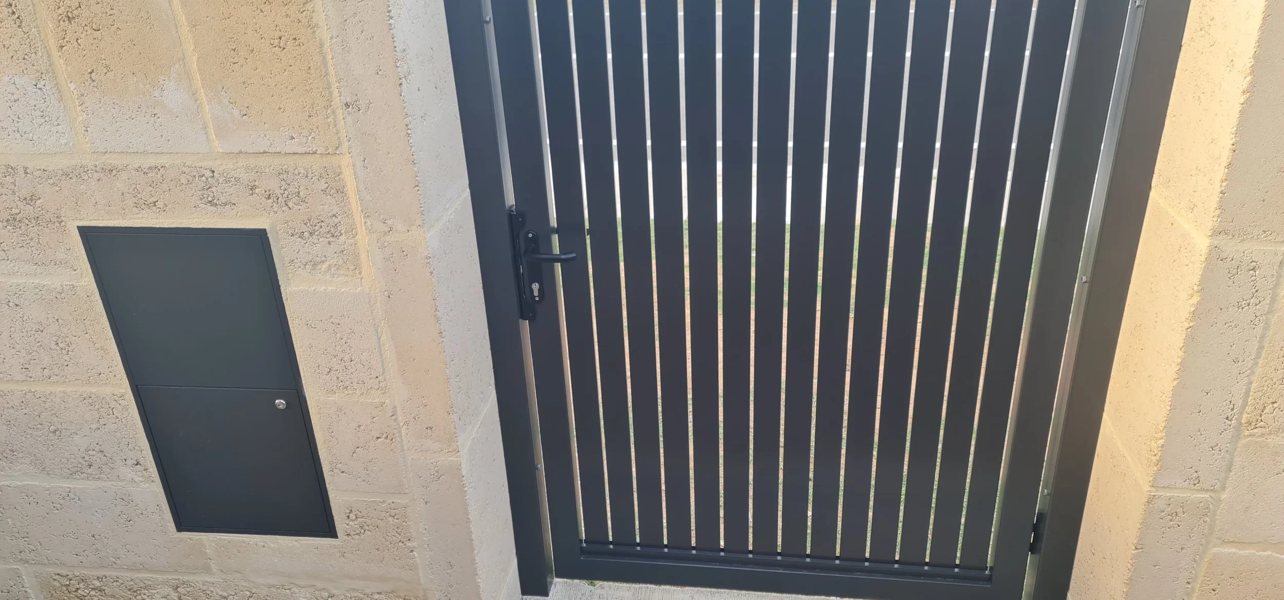 A monument steel gate with vertical slats, attached to a limestone wall that has monument parcel box, with some greenery visible through the gaps of the gate.