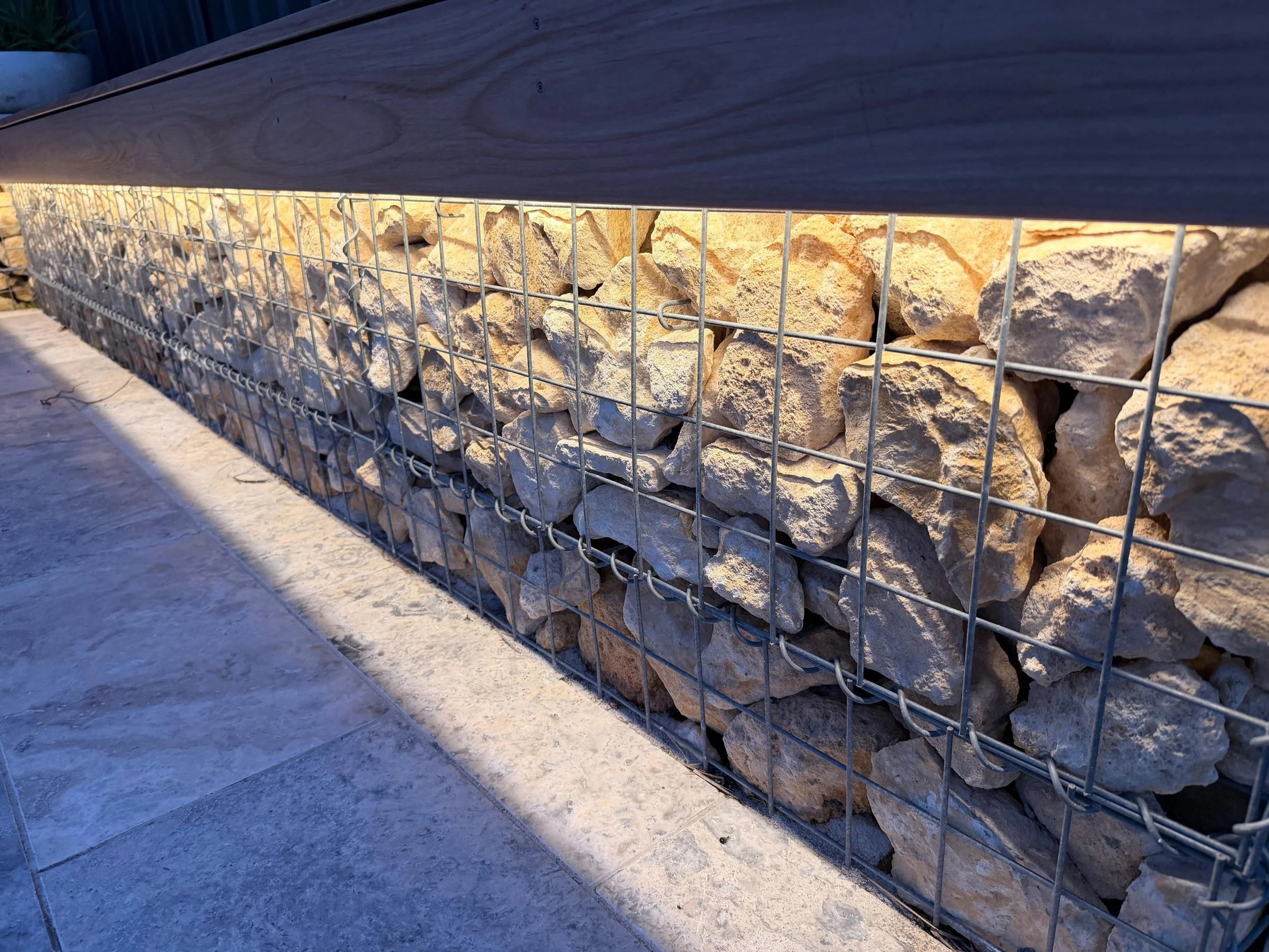 A wire mesh cage filled with large irregularly shaped beige rocks, placed on a smooth outdoor surface, with a wooden panel on top and a potted plant visible in the background.